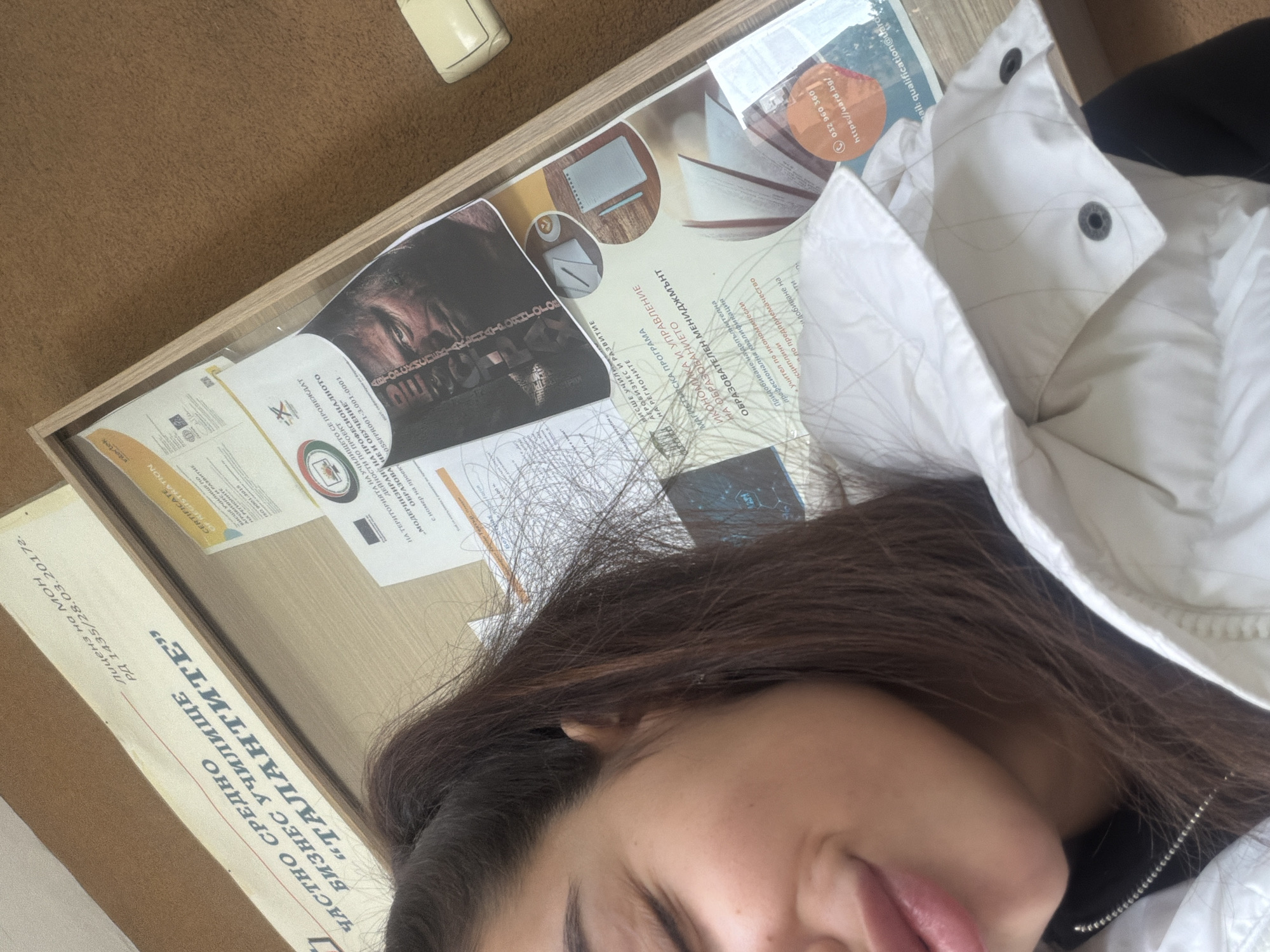 The image shows a close-up of a person's face, partially obscured by a white puffer jacket. The person has brown hair. To the left, there's a wooden frame containing several documents, including certificates and promotional material for "Частно Средно Бизнес Училище "ТАЛАНТИТЕ" (Private Secondary Business School "The Talents"), which is licensed by the Ministry of Education. Some of the documents mention "АГРОБИЗНЕС И РАЗВИТИЕ" (Agribusiness and Development), "ИКОНОМИКА И УПРАВЛЕНИЕ НА ОБРАЗОВАНИЕТО" (Economics and Management of Education), and "ОБРАЗОВАТЕЛЕН МЕНИДЖМЪнт" (Educational Management). A phone number "032 960 360" and an email address "mail: qualification@talented.bg" are visible. Plovdiv, Bulgaria is implied by the Bulgarian Cyrillic text on the documents.