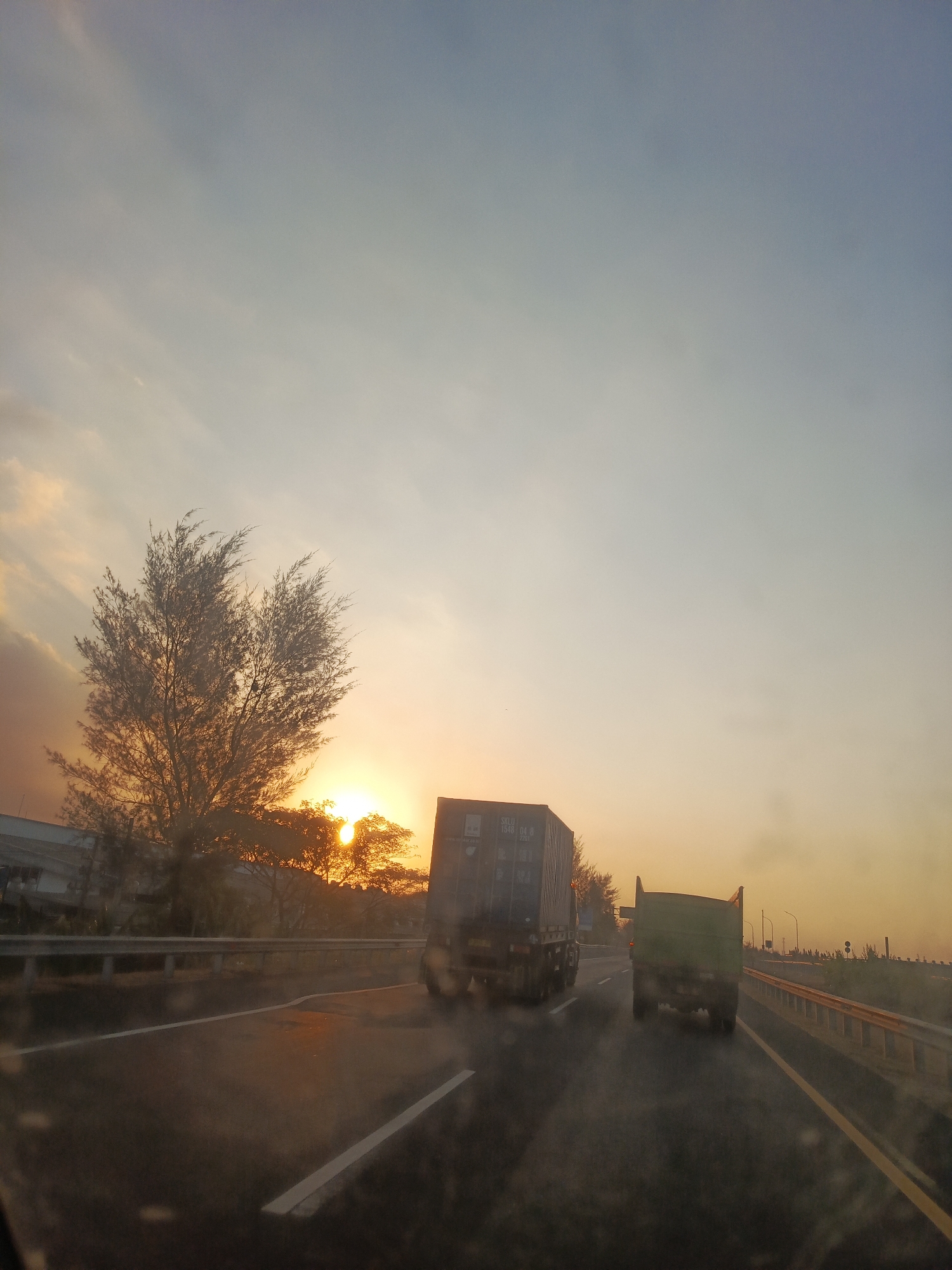 The image depicts a highway scene at sunset, likely in the vicinity of Wonokoyo, Indonesia. The sky is a gradient of soft blues and oranges, with the sun low on the horizon, partially obscured by trees. Two large trucks are visible on the highway, traveling in the same direction. The truck on the left is a container truck, dark in color, with shipping information visible on its side. The truck on the right is a green flatbed truck. Both trucks are in motion, suggesting they are part of the evening traffic. The highway has multiple lanes, indicated by white lines, and a yellow line marking the center. Guardrails line the side of the road. Vegetation, including trees and bushes, frames the highway on the left and right. In the distance, some industrial buildings or structures can be seen on the left side of the road. The overall atmosphere is that of a typical highway commute during a scenic sunset.