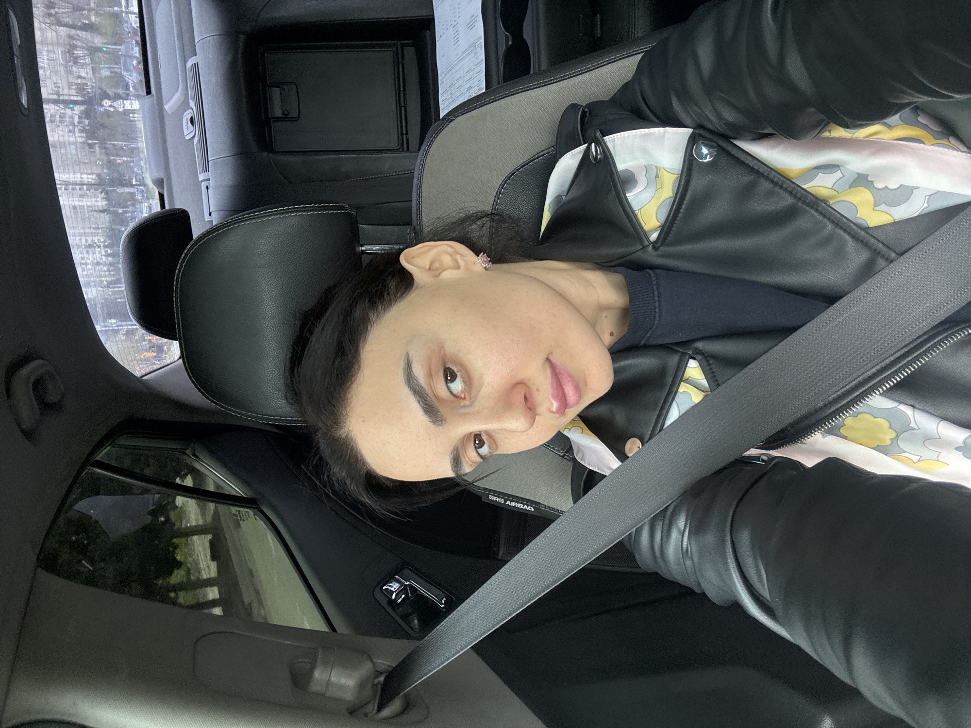 A woman is depicted inside a car, likely in the passenger seat, taking a selfie or being photographed. The image is rotated 90 degrees counter-clockwise. She has dark hair, dark eyebrows, and brown eyes, looking towards the camera with a neutral to slightly contemplative expression. She is wearing a black leather jacket over a light-colored, patterned shirt (featuring yellow, grey, and white cloud-like or wavy motifs) and a dark blue long-sleeved top underneath. A small, possibly pink or purple, stud earring is visible in her right ear. Her lips are adorned with light pink or nude lipstick.
She is securely buckled in with a grey seatbelt crossing her chest and lap. The car's interior is predominantly dark, with black and grey accents. A headrest is visible behind her, and another in the background. The text "SRS AIRBAG" is visible on the side of the car seat. Through the car windows, a blurred urban landscape can be seen, with hints of buildings and trees, suggesting a city environment. The lighting appears soft and diffused, indicating an overcast day. A piece of paper, possibly a document or receipt, is seen in an overhead compartment of the car. The setting is Tbilisi, Georgia.