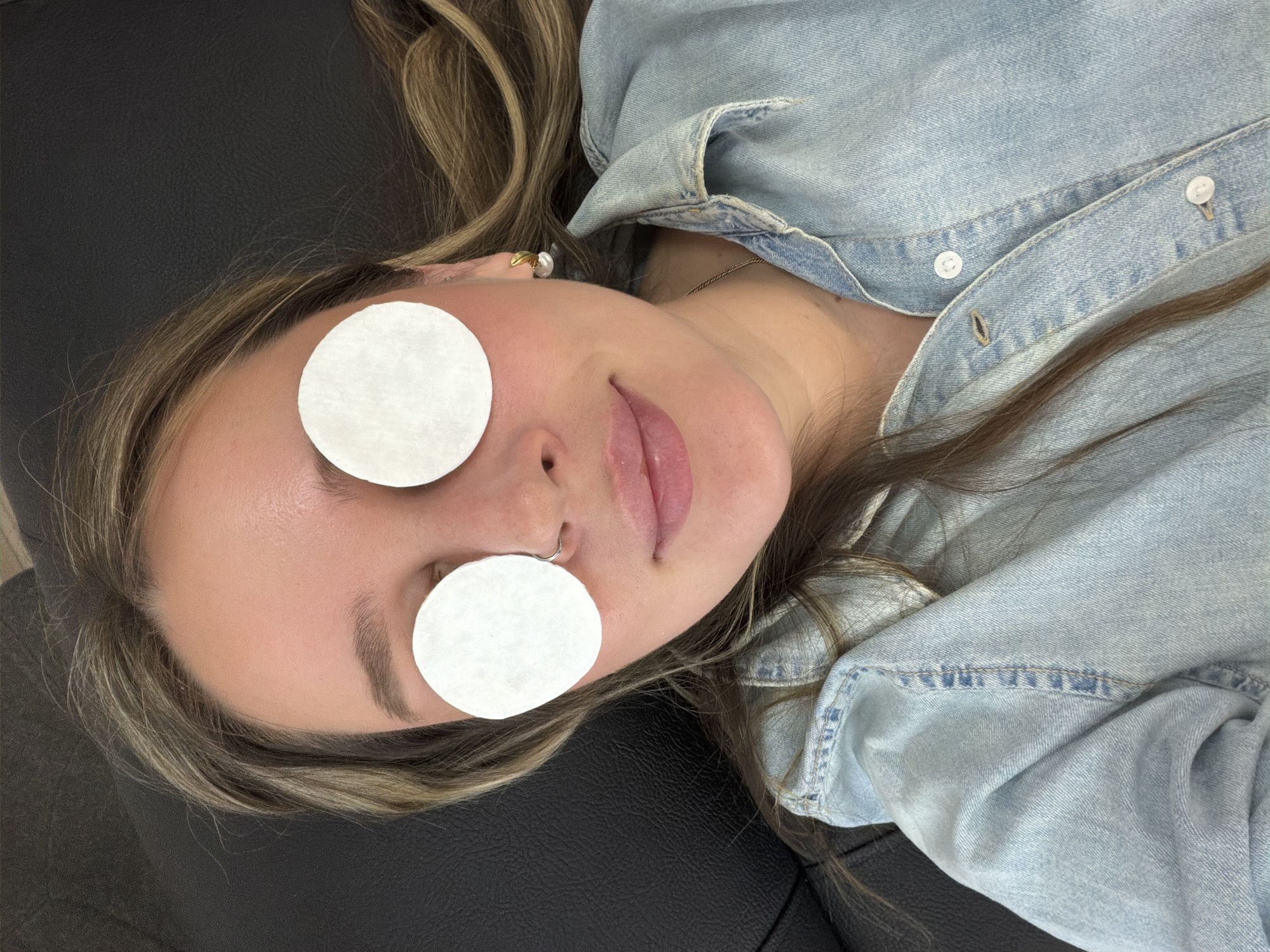 A woman with light brown and blonde hair lies on a dark surface, possibly a couch or table. She is wearing a light blue denim shirt. Two round cotton pads cover her eyes. She appears to be relaxing or receiving some kind of beauty treatment. A gold earring is visible on her right ear. A thin gold chain necklace is around her neck. The location, Chaikovsky, Russia, cannot be determined from the image.