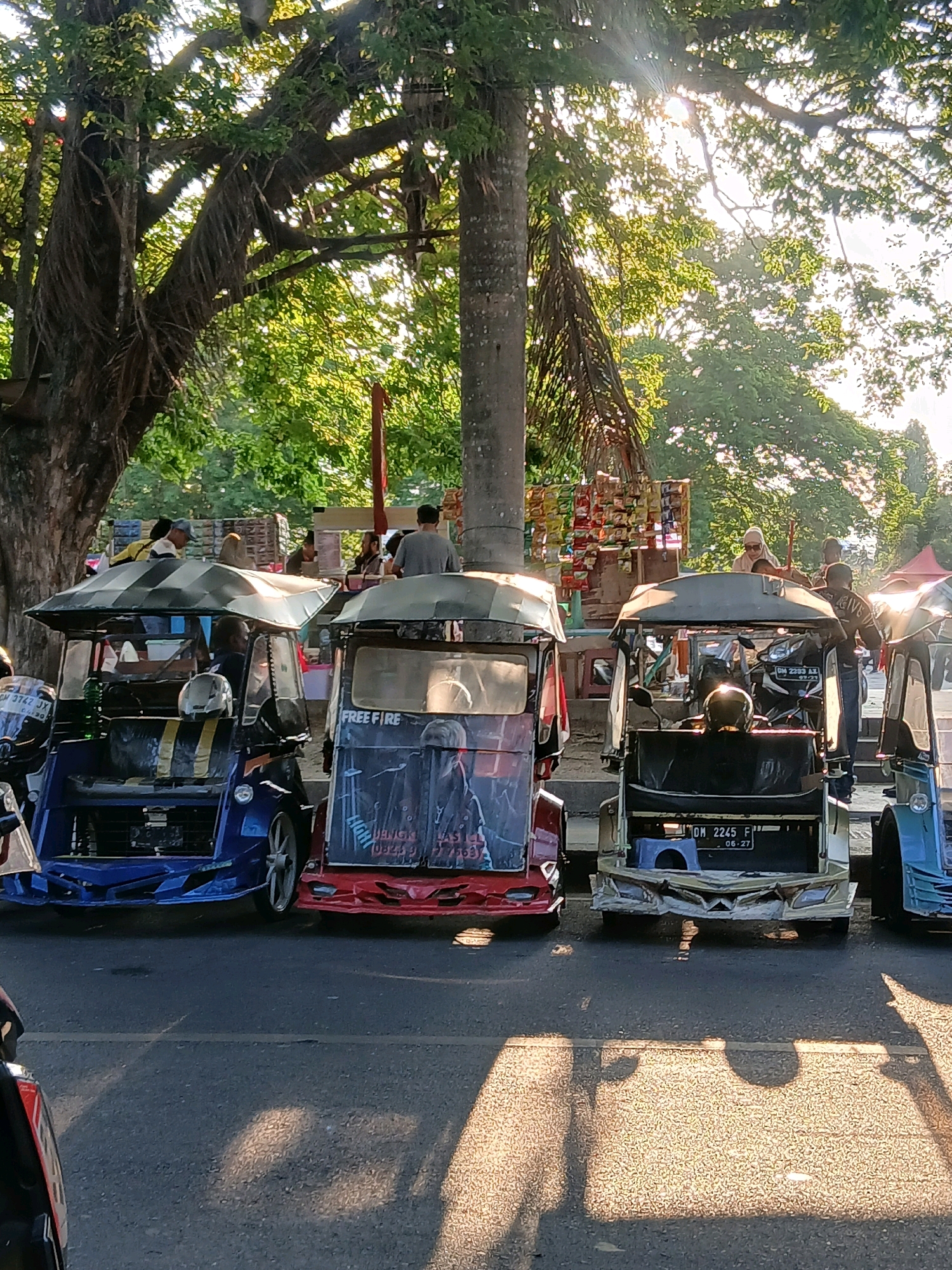 The image depicts a street scene in Gorontalo, Indonesia, where several three-wheeled motorized vehicles, commonly known as "bentor," are parked. The bentors are decorated with colorful graphics and some have canvas awnings. In the background, lush green trees are visible, with sunlight filtering through the leaves, suggesting it is either morning or late afternoon. Several people are present, some standing around the vehicles and others in the background, possibly interacting with vendors. The overall impression is of a bustling local market or transportation hub.