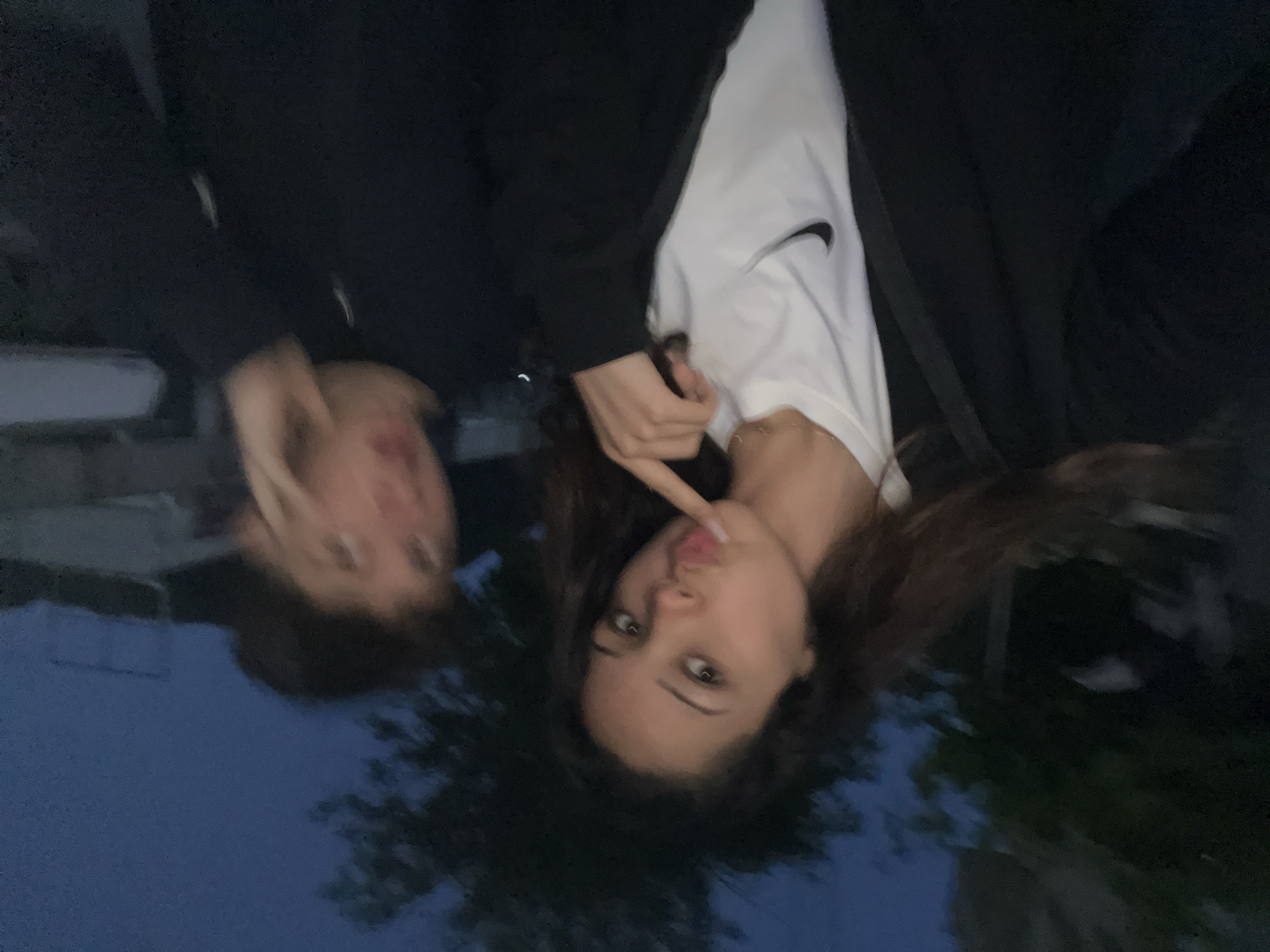 The image depicts two individuals outdoors in Zlatograd, Bulgaria, under low-light conditions consistent with evening or night. On the left, a female subject with long dark hair and light-colored eyes looks directly at the camera. She wears a white t-shirt featuring a black Nike swoosh logo and a dark outer jacket. Her left hand is raised, with the index finger touching her puckered lips. A thin necklace is visible around her neck. To her right, a male subject is present, heavily blurred by motion, wearing a dark long-sleeved garment. His right hand is positioned near his face, also blurred. The background consists of dark, indistinct tree foliage and outlines against a deep blue sky.