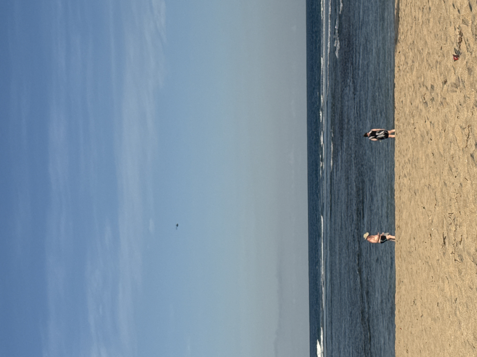On a sunny day, two people are standing on a sandy beach in Kuta Selatan, Indonesia, facing the ocean. The person on the left is wearing a hat and dark shorts, with their back to the camera. The person on the right is wearing a black and white striped dress with black shorts, also with their back to the camera. The ocean is a deep blue with white waves breaking on the shore. A faint wisp of clouds is visible in the sky, and a small helicopter can be seen flying in the distance. The sand is light brown and shows some scattered debris.