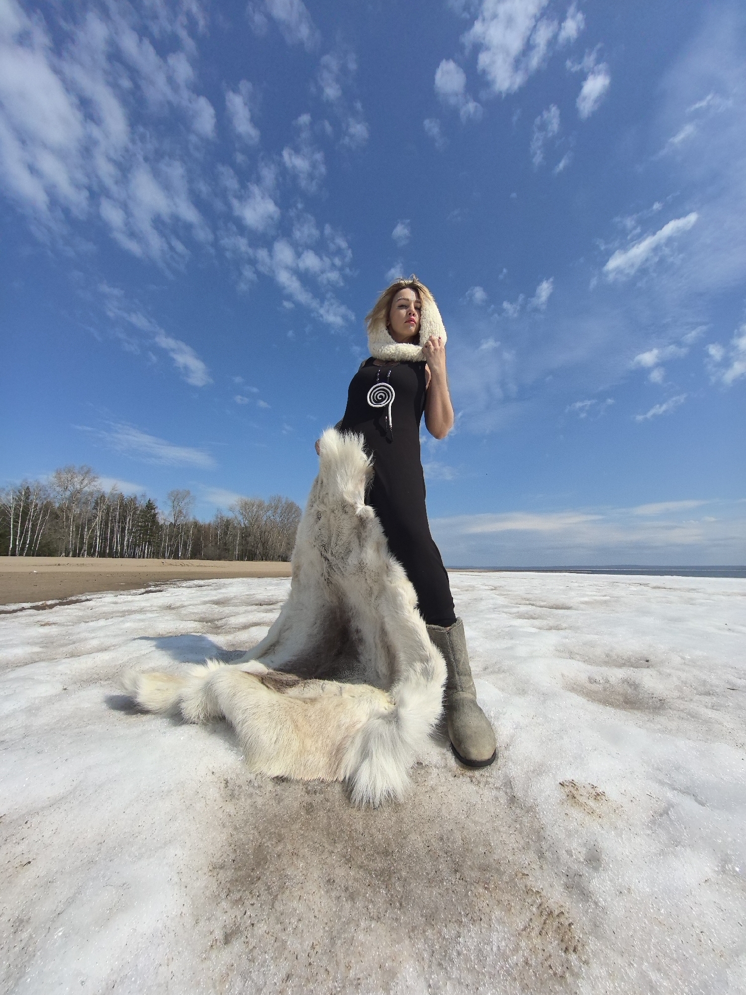A low-angle, wide shot captures a woman posing on a landscape of melting snow and ice under a vast, bright blue sky dotted with scattered white clouds. The woman stands facing slightly right, looking off-camera with a serious or contemplative expression. She has blonde hair and is dressed in a dark, likely black, form-fitting garment, grey boots, and a prominent white furry hood that frames her face. A large, light-colored furry pelt or cape is draped over her left arm, trailing onto the slushy ground beside her.
The foreground is dominated by a mixture of glistening ice, slushy snow, and patches of dark, exposed earth or sand, indicating an early spring or late winter setting. In the background, a body of water, likely a lake or sea, extends horizontally. Beyond the water, a dense forest of tall, bare-branched trees, possibly birches, stretches across the horizon.
Notable details include a white spiral design visible on her black top. The bright sunlight suggests it is daytime, likely late morning or early afternoon. The stark yet beautiful natural environment, with its mix of snow, water, and forest, aligns with the provided location context of Noviy, Russia.