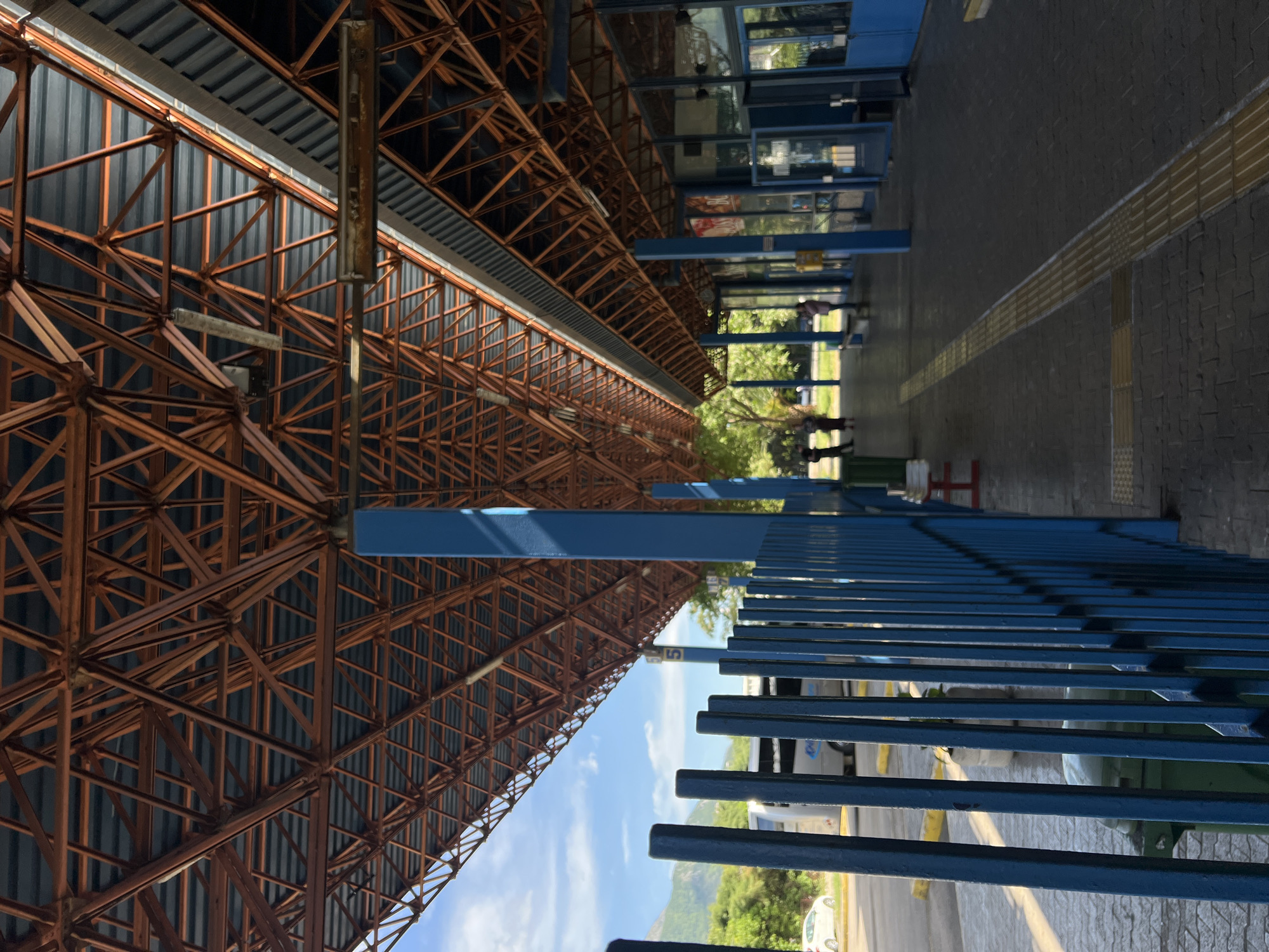 The image captures an expansive, outdoor public transport station or sheltered area, likely located in Bjeliši, Montenegro, under bright daylight. A dominant feature is a massive, rust-colored or dark orange metal truss structure forming an extensive canopy or roof overhead. This intricate framework of interlocking triangular beams supports a roof made of dark grey corrugated panels. Several prominent blue columns provide additional support for the structure.
Parallel to the roof, on the right side of the frame, runs a long, blue metal fence or railing, designed to guide pedestrians or separate areas within the station. The ground beneath is paved with dark grey interlocking bricks, featuring a distinctive yellow tactile paving strip that stretches into the background, intended to assist the visually impaired.
In the mid-ground and background, beyond the blue fence and some smaller blue structures (possibly kiosks or bus shelters), a few blurry human figures are visible. One figure appears to be pushing a cart. The faint outlines of buses or other large vehicles can be seen further back, affirming the setting as a transportation hub. A small white car is also partially visible in the far distance.
The sky is clear and blue with scattered white clouds, indicating sunny weather. Lush green foliage from trees and bushes frames the background, and the gentle contours of hills or mountains are discernible on the horizon. A white sign with the black number "5" is visible in the distance, likely indicating a bus bay or platform. The scene depicts a functional and well-structured public transport facility.