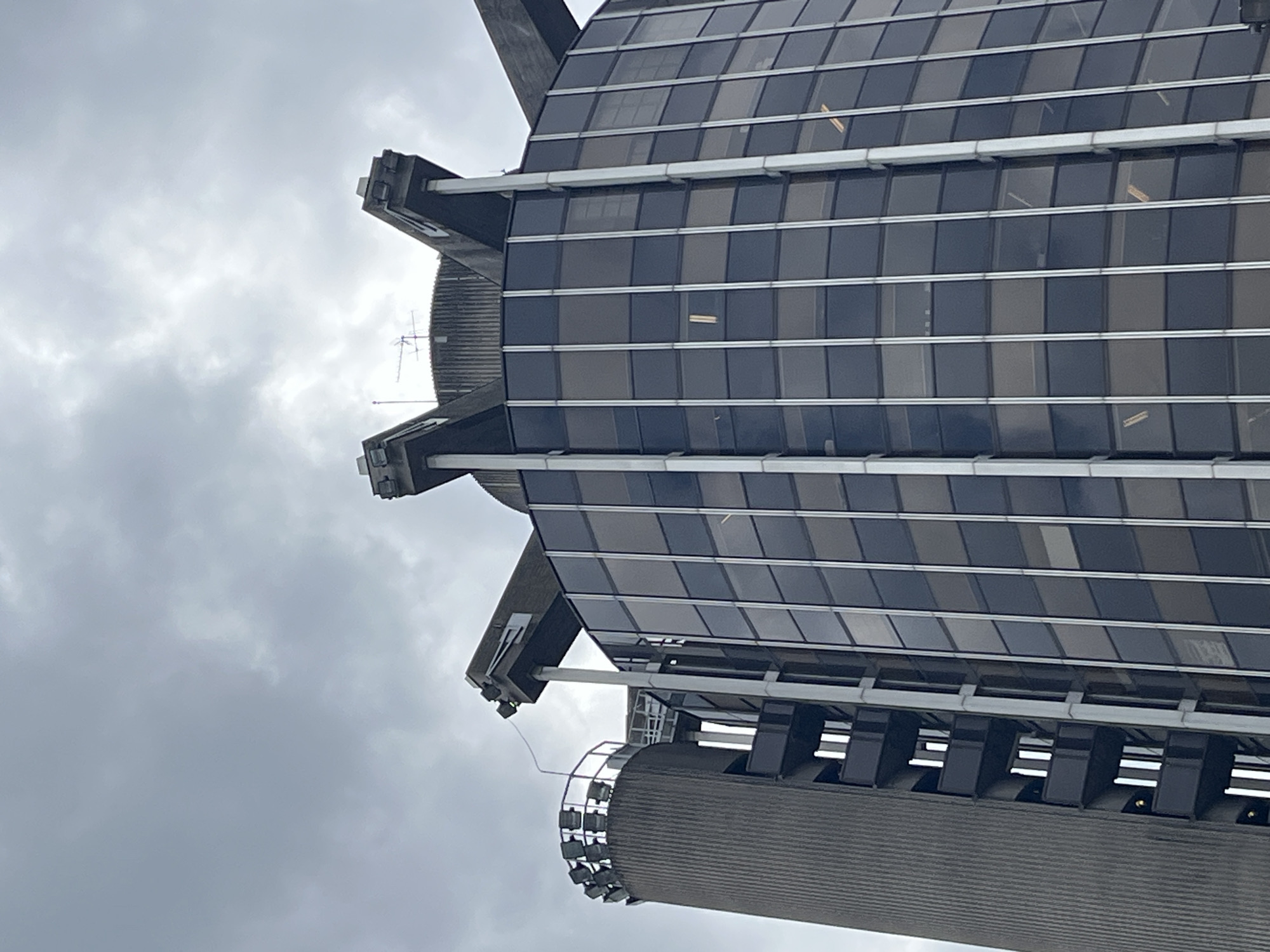 The image is a low-angle, vertically oriented shot of a modern building with a glass facade, set against an overcast sky. The building's architecture is characterized by sharp, angular protrusions and horizontal bands of windows. Some windows appear to be lit from within, suggesting it's either daytime with interior lighting or dusk/night. The building has a ribbed cylindrical section at the bottom, topped with a ring of spotlights. The sky is filled with dense, grey clouds, indicating a cloudy or possibly rainy day. There is no visible text in the image. The scene appears to be an urban environment, likely in Créteil, France, given the location context. No people are visible in the frame.