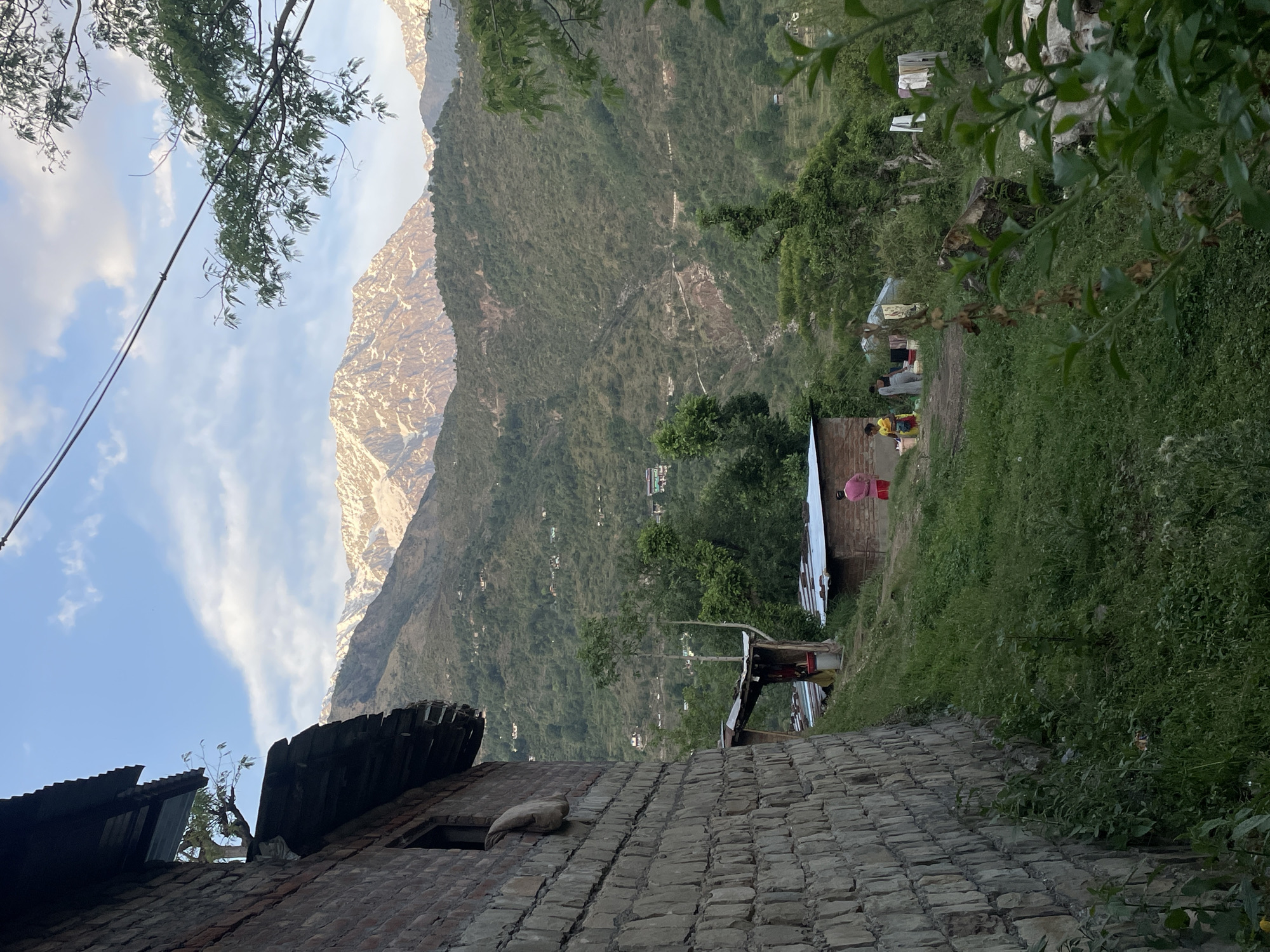 Captured from a low-angle perspective, the image presents a rural scene in Dharamshala, India. The foreground features a brick building with a partially corrugated metal roof. A second, smaller structure is visible, with people gathered nearby. Lush vegetation is prominent, interspersed with glimpses of other buildings and dwellings. In the background, a large, mountainous landscape dominates, with snow-capped peaks under a sky with sparse clouds.