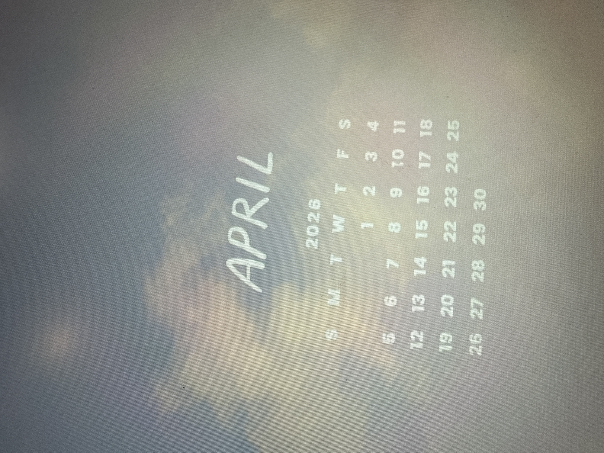 The image shows a calendar for April 2026 with a background of a cloudy sky. The word "APRIL" is printed in large letters on the left side of the calendar. The days of the week are listed as "S M T W T F S" above the dates, which run from April 1st to April 30th. There is nothing to suggest that this calendar is located in Davie, Florida, or anywhere else for that matter. The image does not depict any people, specific objects, actions, interactions, or detailed environment, beyond the calendar itself and the sky background.