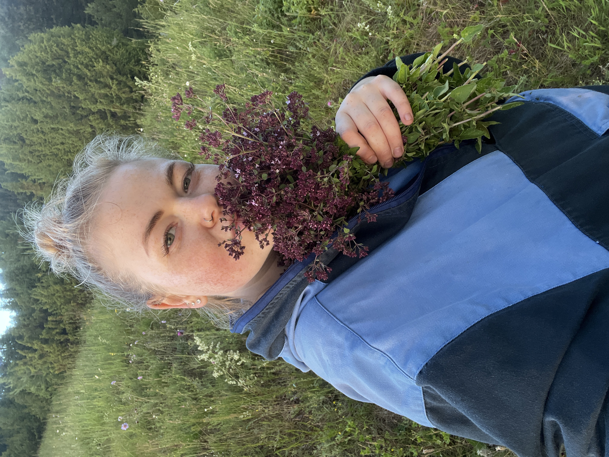 A young woman with blonde hair pulled up in a bun is holding a bunch of wild herbs with purple flowers in front of her face. She is wearing a blue and black jacket. The woman is in a grassy field with trees in the background. The location is the city of Olkhovka, Russia.