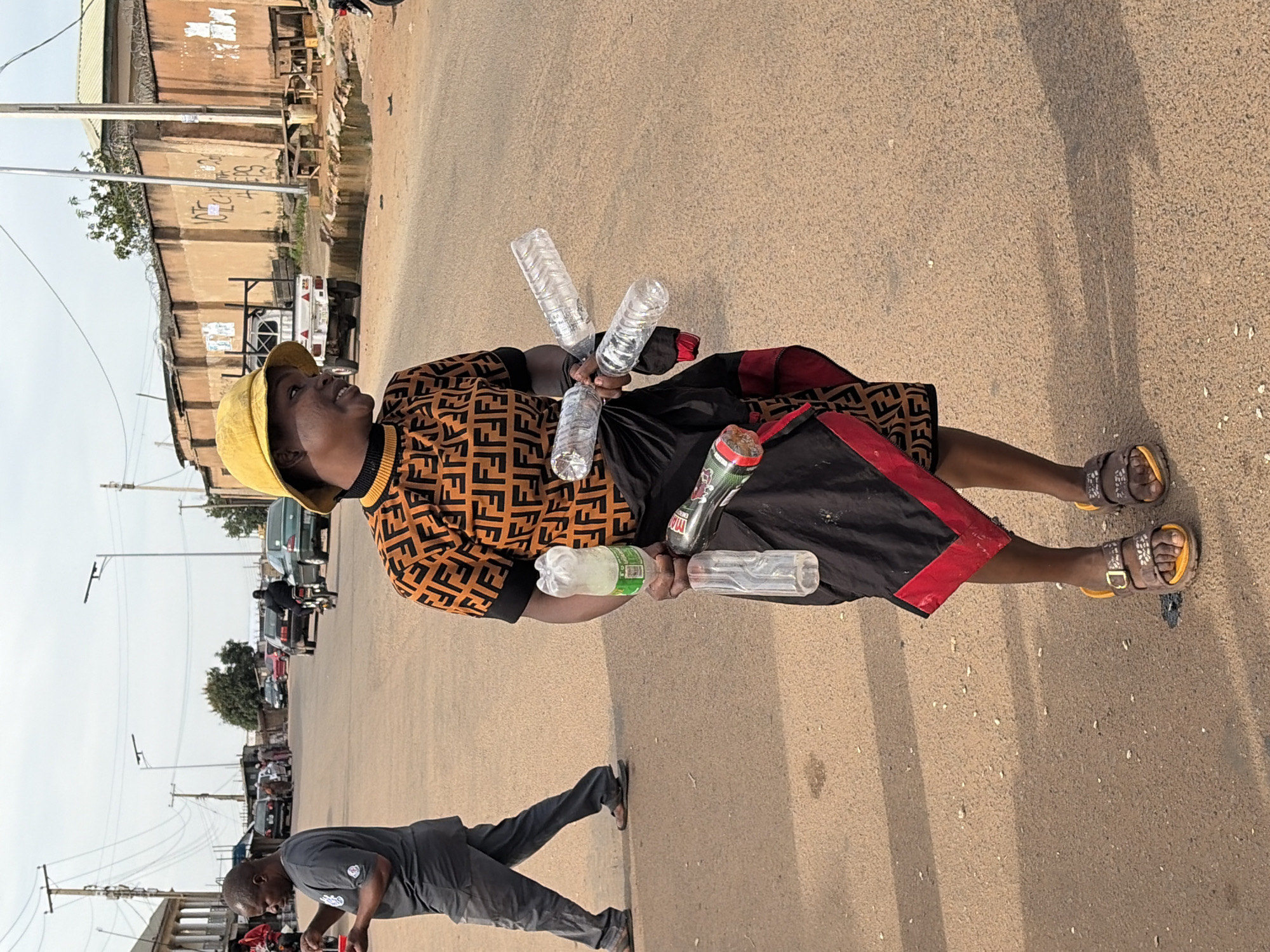 The image depicts a street scene in Gura Top, Nigeria, where a person wearing a yellow bucket hat, an orange and black patterned shirt, and a black skirt with a red trim is holding several plastic bottles and a red-capped object. The individual is standing on a dirt road with other people and vehicles in the background. To the left, a man in a gray t-shirt and pants walks away from the camera. Several cars and a motorcycle are visible on the road, and buildings line the sides of the street. The environment appears to be an urban or semi-urban setting with overhead power lines and some trees. The lighting suggests it is daytime.