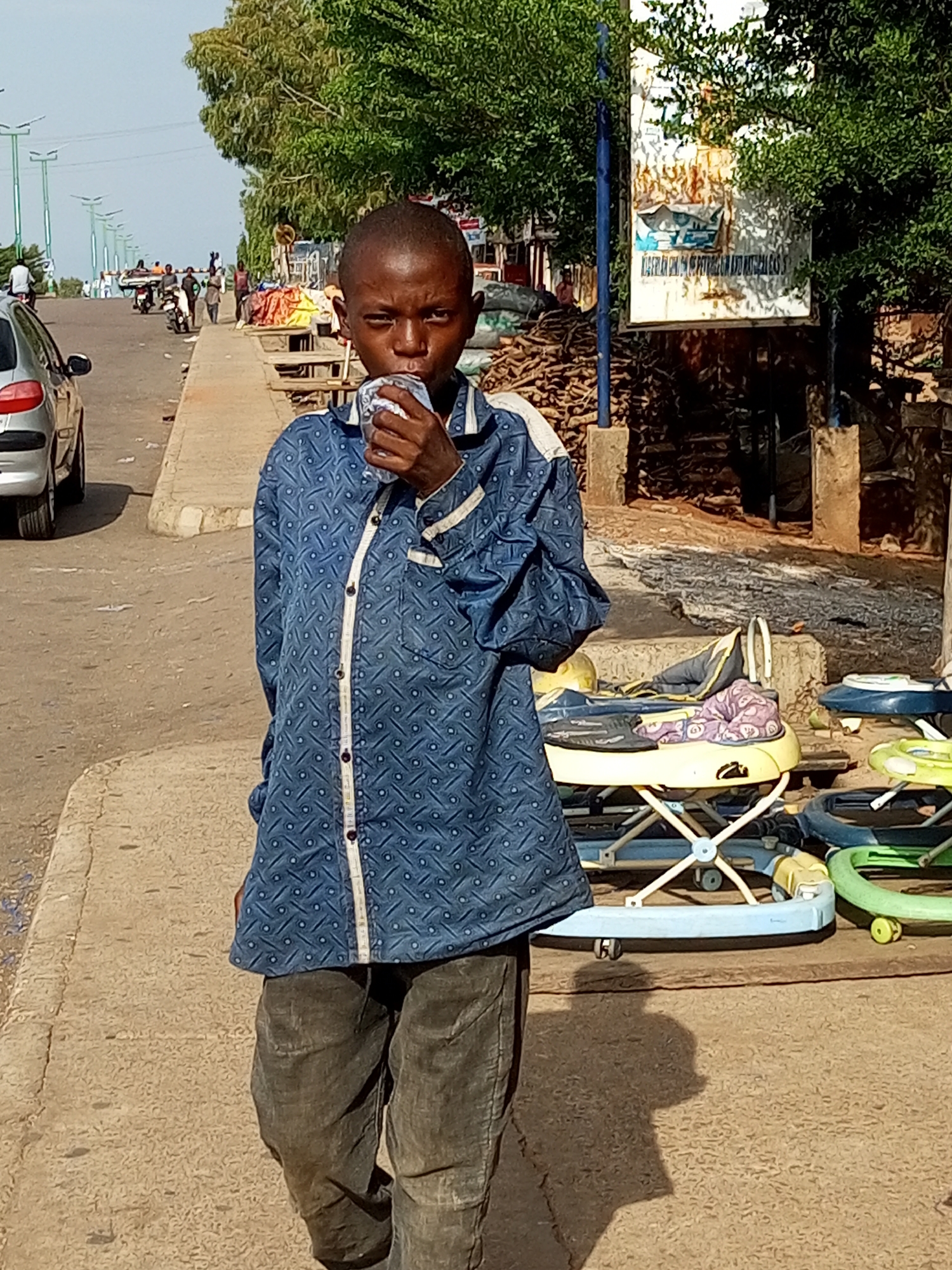 A young boy stands on a sidewalk, holding a drink to his mouth. He is wearing a blue patterned shirt and dark pants. In the background, a street is visible with cars and people walking. There are also some stalls with goods for sale. A baby walker is visible in the foreground, suggesting a residential or market area. The lighting suggests it is daytime, possibly morning or afternoon. The overall scene depicts a typical street in Gombe, Nigeria, with everyday activities taking place.
