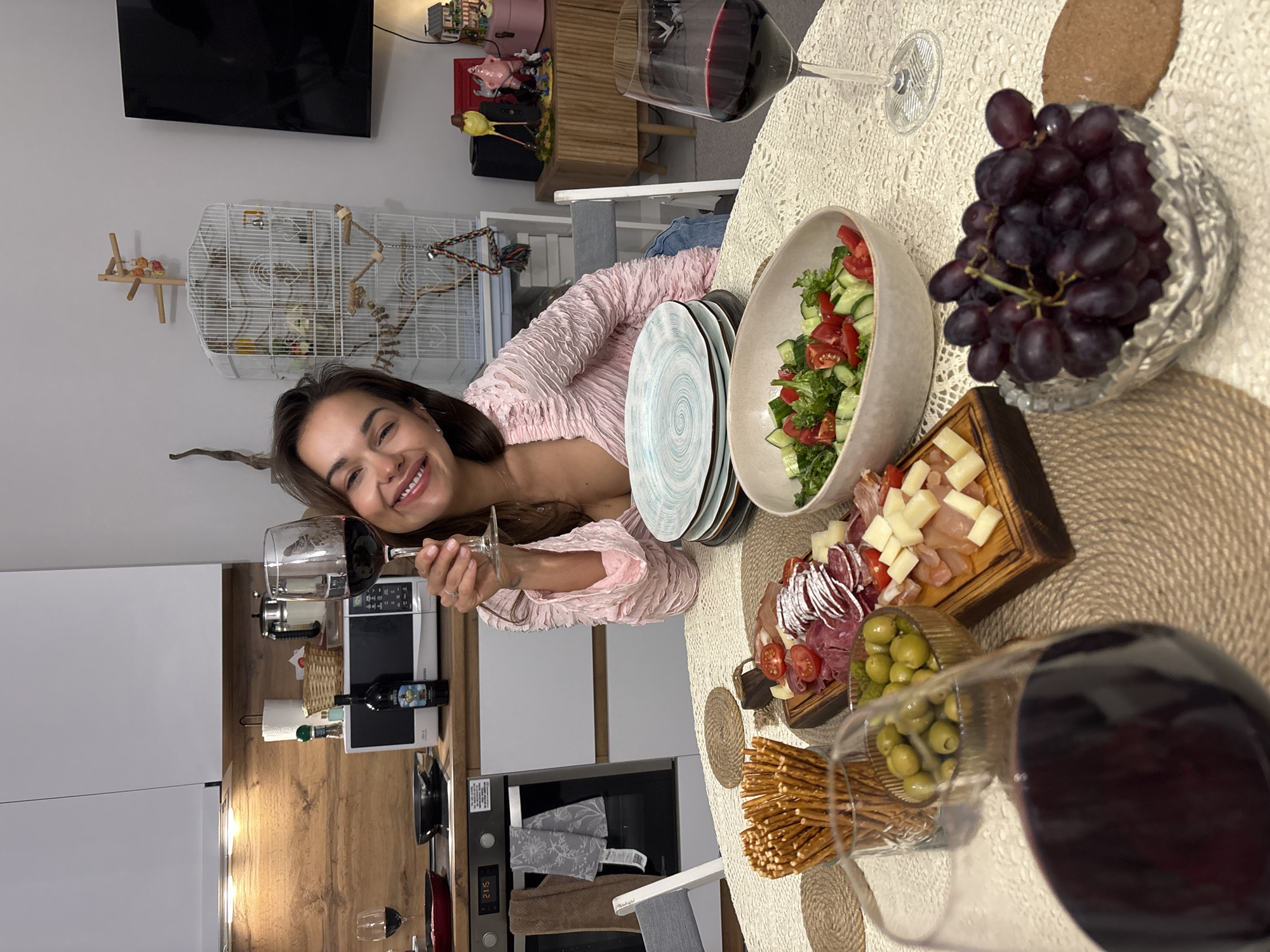 The image, captured in Krasnoyarsk, Russia, shows a young woman smiling and holding a glass of red wine, indicating a relaxed and enjoyable atmosphere. She has long dark hair and is wearing a light pink, textured top.
The foreground is dominated by a table set with a variety of food and drinks. Visible items include a bowl of fresh green salad with tomatoes and cucumbers, a crystal bowl brimming with dark grapes, a charcuterie board laden with sliced meats (such as salami) and cubed cheeses, a glass container of green olives, and a bowl of pretzel sticks. Several glasses of red wine are present on the table, which is covered with a light-colored, lacy tablecloth and features braided placemats. A stack of patterned plates is also visible.
The background reveals a modern home interior, combining a dining area with a kitchen. The kitchen features white cabinets, wooden countertops, and various appliances, including a microwave and an oven, whose digital display reads "21:15". A wine bottle, partially labeled "MONTEPULCIANO D'ABRUZZO," is on the kitchen counter. Above, a wall-mounted television is visible, along with decorative figurines (including a pink pig and a yellow bird) on a shelf. To the left, a white bird cage houses two small pet birds, adding a domestic touch to the scene. The woman's expression of happiness suggests a pleasant social occasion or meal.