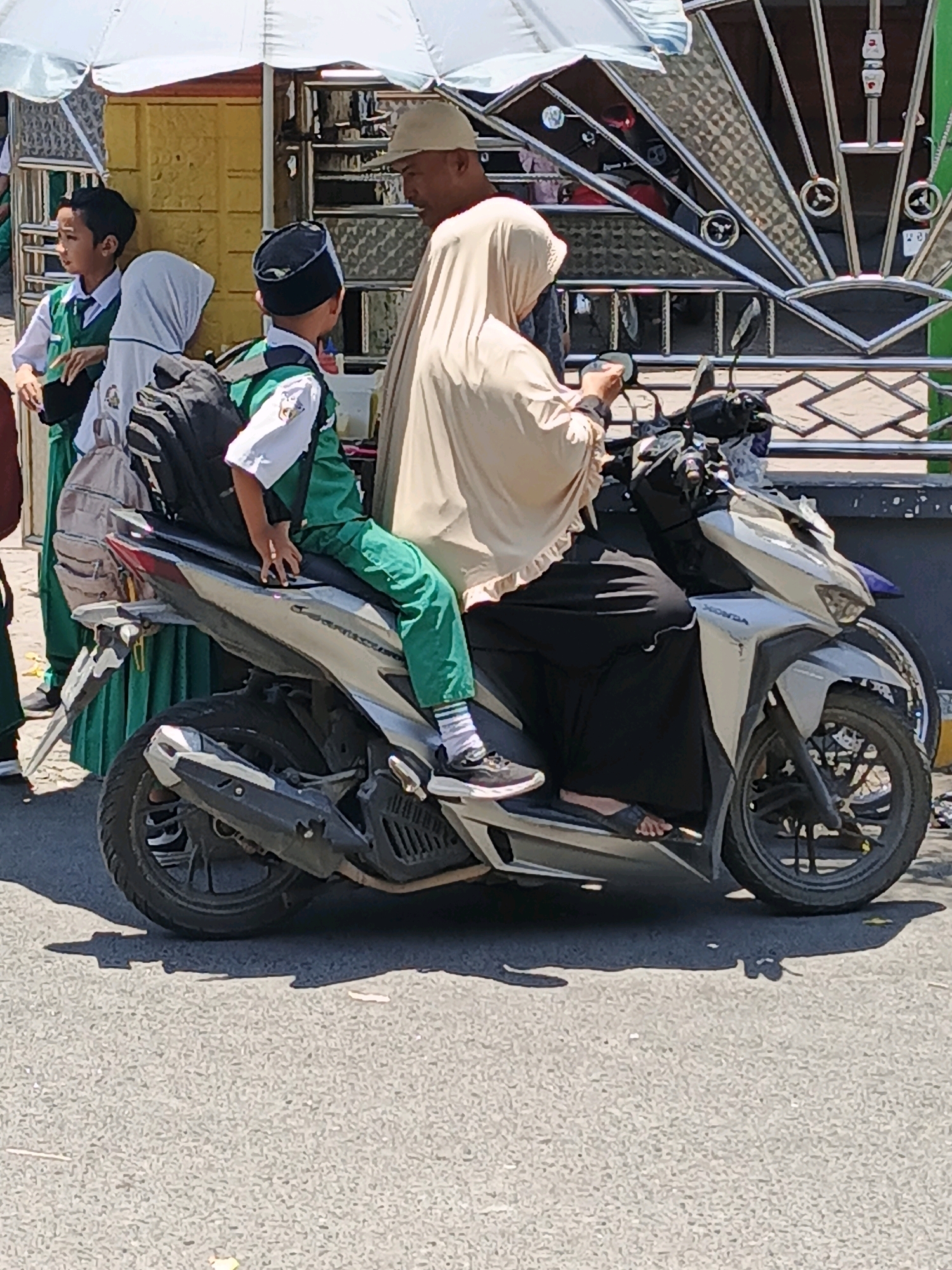 In Ketegan, Indonesia, a young boy in a green school uniform, complete with a white shirt, vest, and green pants, sits on the back of a silver Honda scooter. He wears a black cap and white socks with sneakers. The scooter is driven by a woman wearing a light brown hijab and a black abaya, with her feet in black sandals. They appear to be stopped, possibly waiting. Beside them, another child in a green uniform and white hijab is visible, as well as a man wearing a light-colored hat, who is turned towards the scooter. The background shows a yellow structure with metal detailing and an umbrella providing shade. The ground is asphalt.