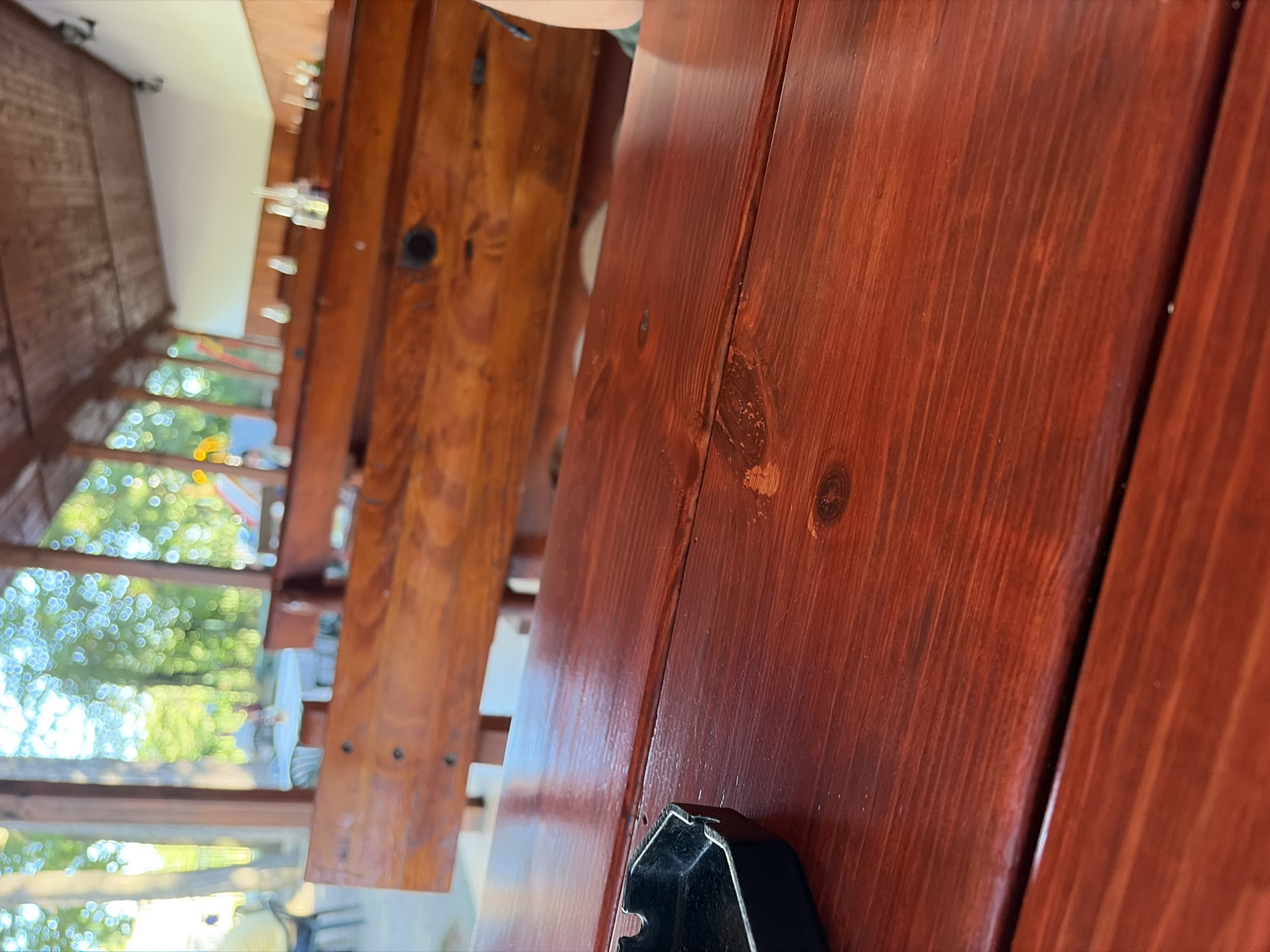 The image shows an outdoor seating area with wooden tables and benches. In the foreground, a reddish-brown wooden table has an ashtray on the left side. In the background, rows of wooden benches are visible, with a blurry view of trees and greenery through a covered area. The overall impression is of a casual, outdoor dining or relaxation spot. No specific details about the city of Koprinka, Bulgaria are visible in the image itself, and the scene does not provide any information that could link it to a specific city or location.