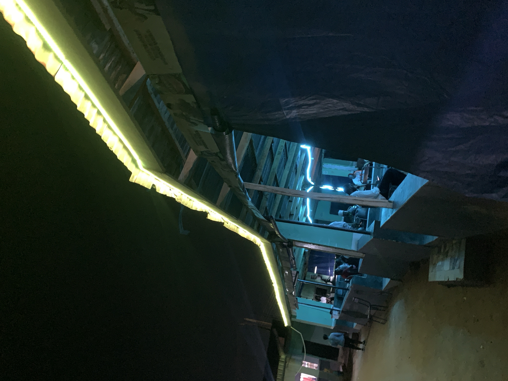 At night near New Stadium in Nigeria, a poorly lit outdoor area is covered by a dark tarp strung beneath a partially illuminated building, possibly a market or series of shops. The exterior of the building is lined with yellow LED strip lighting. A single person walks on an unpaved ground. The area is sparsely furnished with metal chairs. Through gaps in the tarp, an interior lit with blue lighting is visible, with numerous other people inside the building.