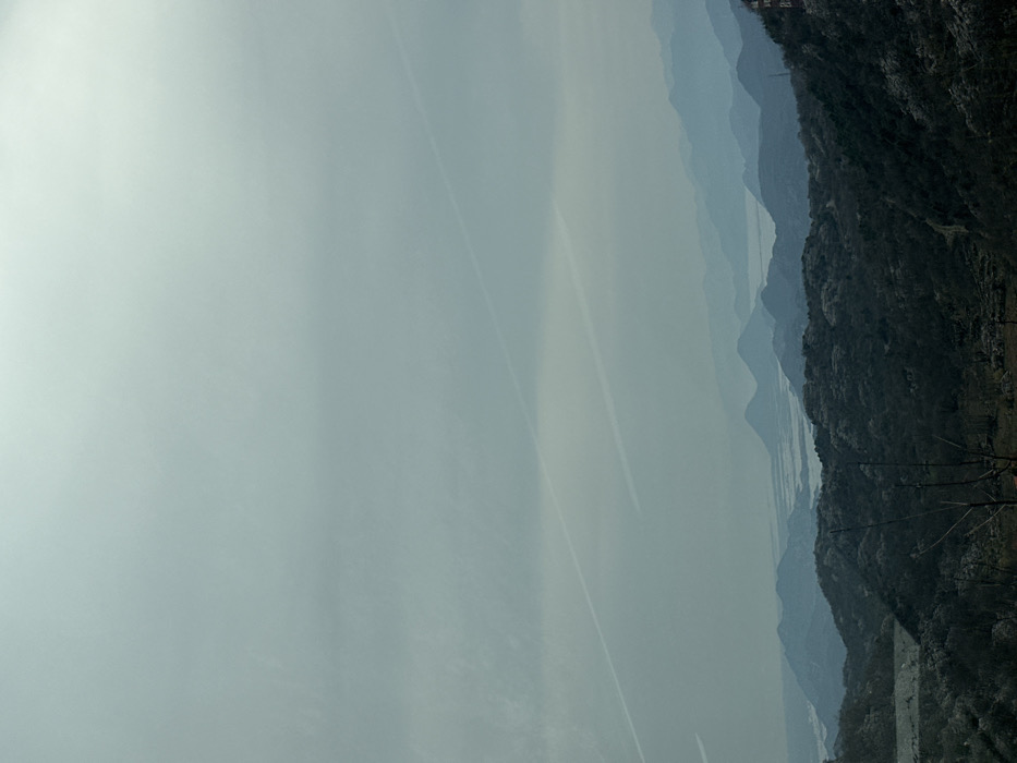 This image presents an expansive landscape vista observed from an elevated position in the area of Gornji Ulići, Montenegro. The scene is dominated by a largely overcast, light grey sky that occupies the upper two-thirds of the frame. Two distinct, parallel white contrails are visible within the sky, traversing diagonally from the upper left quadrant towards the lower right. Below the sky, multiple layers of mountains and hills recede into the distant haze, displaying a gradual blue tonal shift indicative of atmospheric perspective. A large body of water, consistent with a lake or wetland system, is visible horizontally across the midground, situated between the distant mountain ranges. The immediate foreground features dark, densely vegetated hillsides composed primarily of bare or sparsely leafed trees, suggesting a dormant or early growth season. A small, reddish-brown structure is partially visible on a distant hillside at the far right of the image. No individuals, specific actions, or direct interactions are present.