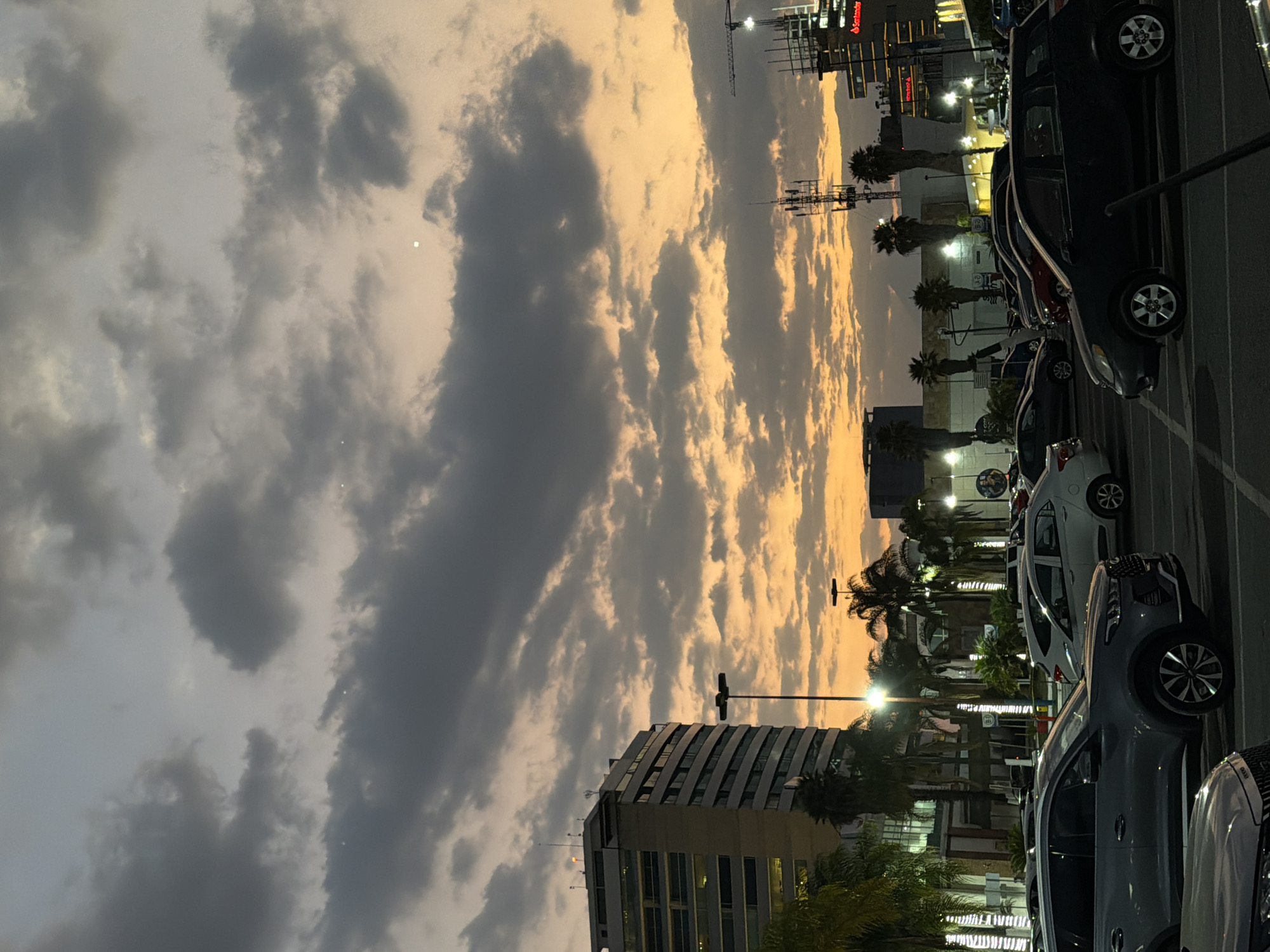 The image depicts an urban commercial area at dusk in San Bernardino Tlaxcalancingo, Puebla, Mexico. The sky is largely covered by dense, textured clouds, which display warm orange and yellow gradients along the horizon, transitioning to dark grey and blue-grey overhead. A single bright celestial body is visible in the upper portion of the sky.
In the foreground and midground, a large asphalt parking lot is filled with numerous parked vehicles of various types, including sedans and SUVs. Several tall, dark lamp posts emit bright white light, illuminating segments of the parking area and adjacent structures. On the left, a multi-story building with a modern facade featuring horizontal bands and numerous illuminated windows rises prominently. Clusters of palm trees and other tropical foliage are visible throughout the scene, some decorated with white string lights.
Towards the right mid-ground, a tall cellular communication tower equipped with multiple antennas is present. Further to the right, additional commercial buildings are visible, one featuring an illuminated "Santander" sign. A large construction crane is silhouetted against the sunset on the far right horizon. No individuals are discernible within the frame.