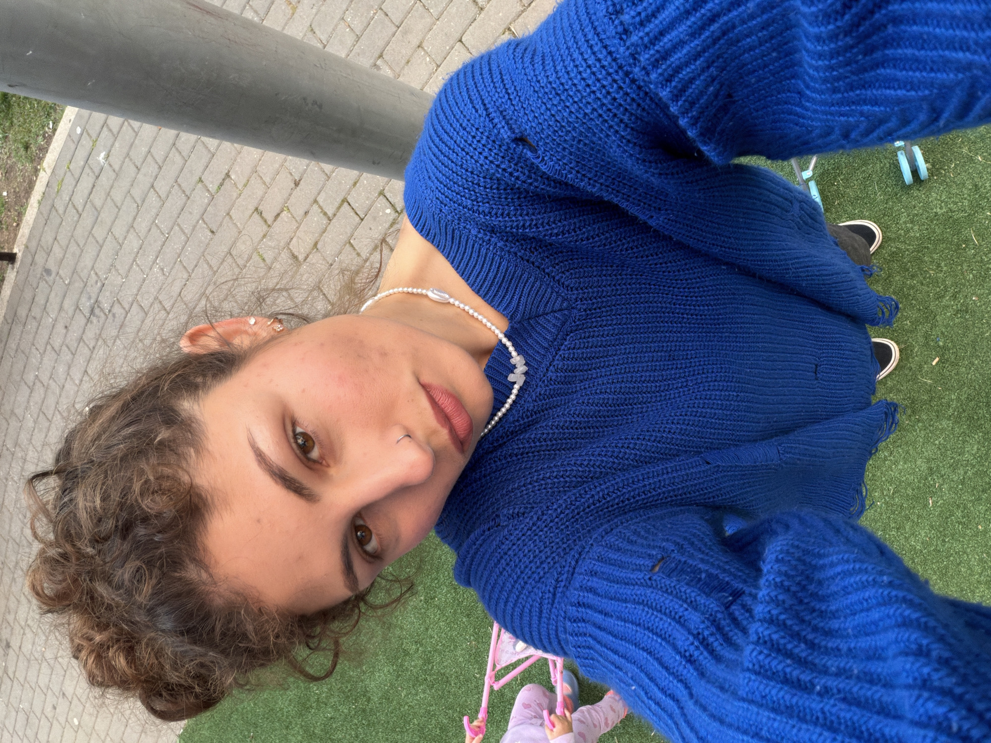 The image is a low-angle selfie featuring a young person with light brown, curly hair and brown eyes, looking directly at the camera. They wear a small nose ring and a pearl necklace with a prominent cowrie shell charm. Small stud earrings are visible in their ear. The person is wearing a vibrant blue knitted sweater, which shows some visible wear or loose threads on the sleeves.
The background is an outdoor setting. A gray metal pole runs diagonally across the upper left. Below this, a paved walkway made of light gray rectangular pavers is visible, bordered by a strip of natural grass. The dominant ground cover in the midground and foreground, especially to the right, is green artificial turf.
On the artificial turf, a child's pink play stroller or pram is partially visible, with a child's hands, dressed in light pink patterned clothing, holding onto its handles. Further back on the turf, a pair of black sneakers with white soles and some light blue plastic objects can be seen. The scene suggests a daytime activity, possibly playing in a park or playground in Kiryat Bialik, Israel. No discernible text is visible in the image.