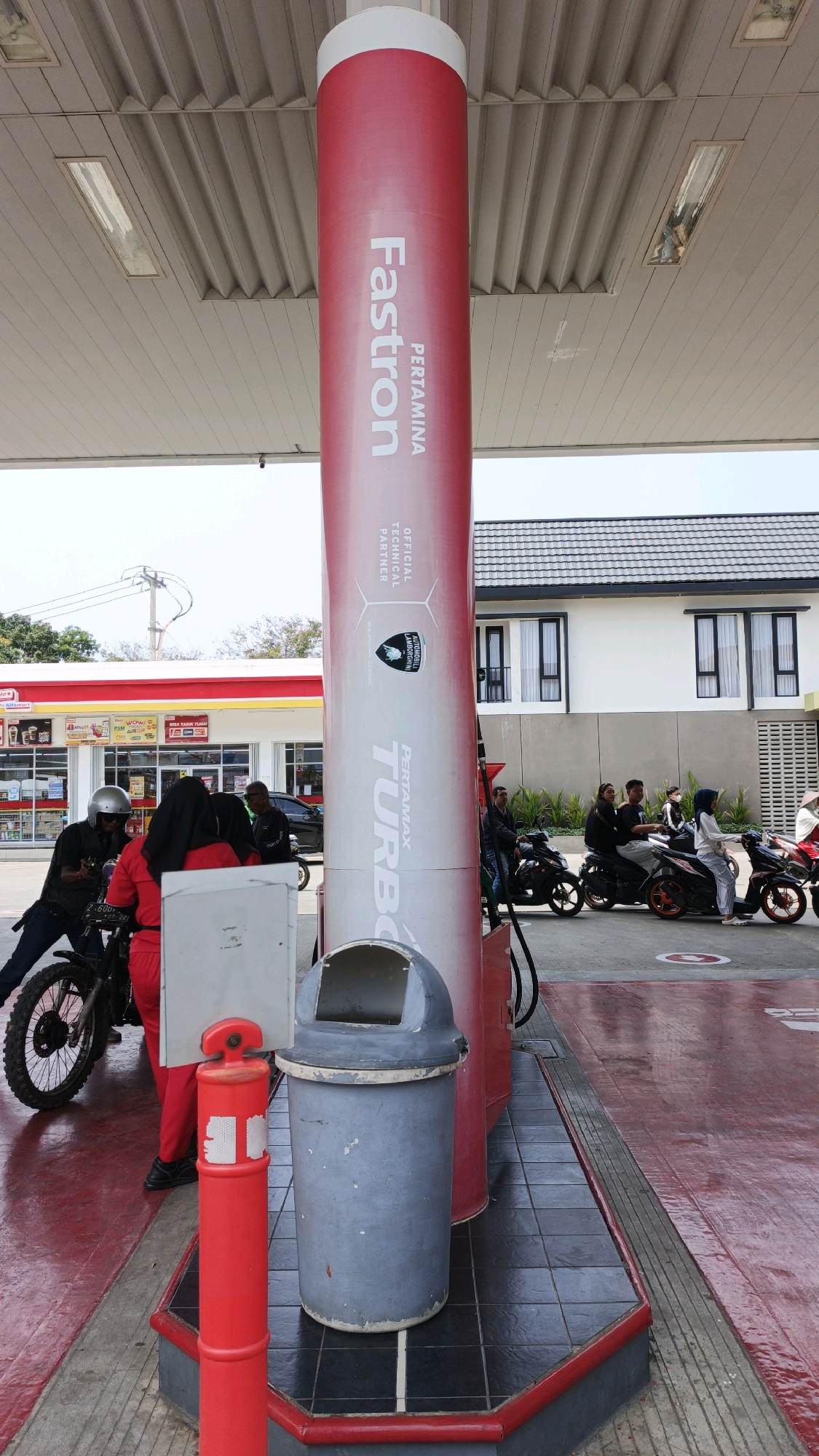 At a Pertamina gas station in Desa Wonoharjo, Indonesia, a large red and white cylindrical fuel pump stands prominently in the foreground. The pump features branding for "Fastron" and "Pertamina Turbo," along with an "Automobili Lamborghini" logo indicating a technical partnership. To the left of the pump, a person wearing a red jumpsuit is near a motorcycle, with another person wearing a helmet and dark clothing also present. Further back, several individuals are seen on motorcycles, some queuing. A blue trash can is situated near the base of the fuel pump. The background shows the canopy of the gas station, a convenience store with red and white awnings, and modern-looking white buildings with dark-framed windows. The ground is a mix of red and grey pavement tiles and a red wet surface. The sky is overcast.