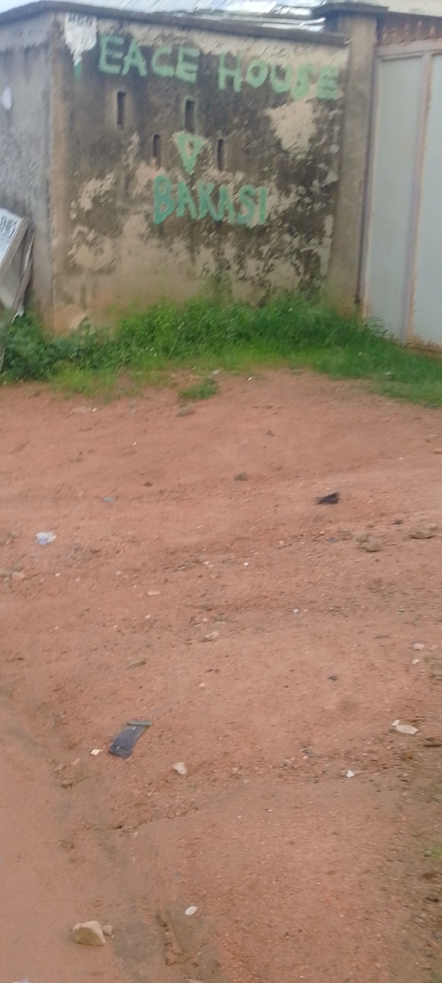 The image shows a concrete wall with "PEACE HOUSE V BAKASI" spray-painted in green on its surface. The wall appears weathered and has patches of dark discoloration. Below the wall, there is a strip of overgrown green grass. The foreground consists of a reddish-brown dirt or sand surface, with a few scattered rocks and debris, including what appears to be a dark rectangular object that could be a piece of electronic equipment or trash. The overall scene suggests an outdoor environment, likely in a less developed area. Without further context or geographical markers, the specific location within Jalingo, Nigeria cannot be determined from the image alone.