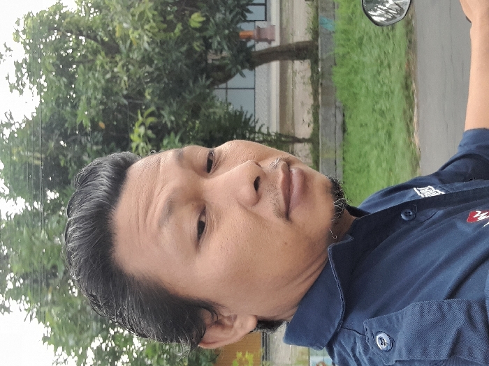 A man with dark hair and a trimmed beard is wearing a blue collared shirt. He appears to be outdoors, potentially on a motorcycle given the presence of a mirror. The background features greenery including trees, and a building is faintly visible. The image appears to be taken from a slightly lower angle. The light suggests it is daytime.