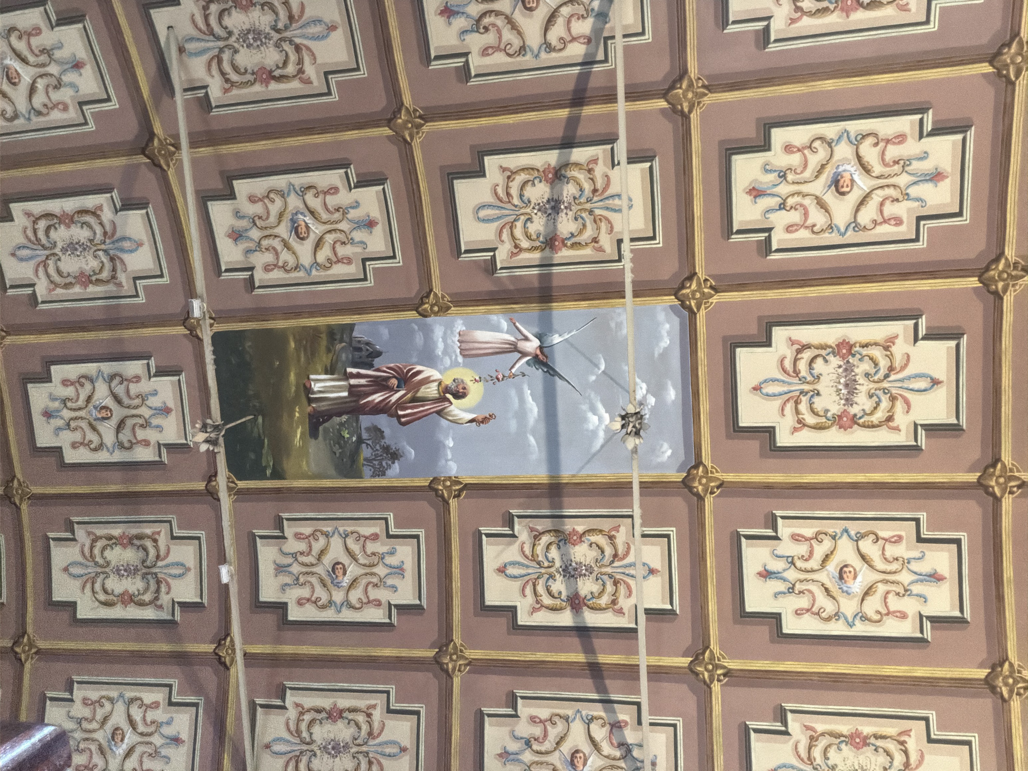 The image shows the decorated ceiling of the Palácio de Mateus, located in Vila Real, Portugal. The ceiling is composed of a grid of rectangular panels, most of which contain decorative floral motifs and angel faces. A larger, central panel depicts a man in robes with a halo and an angel in a sky-like background. Two support beams intersect the view.