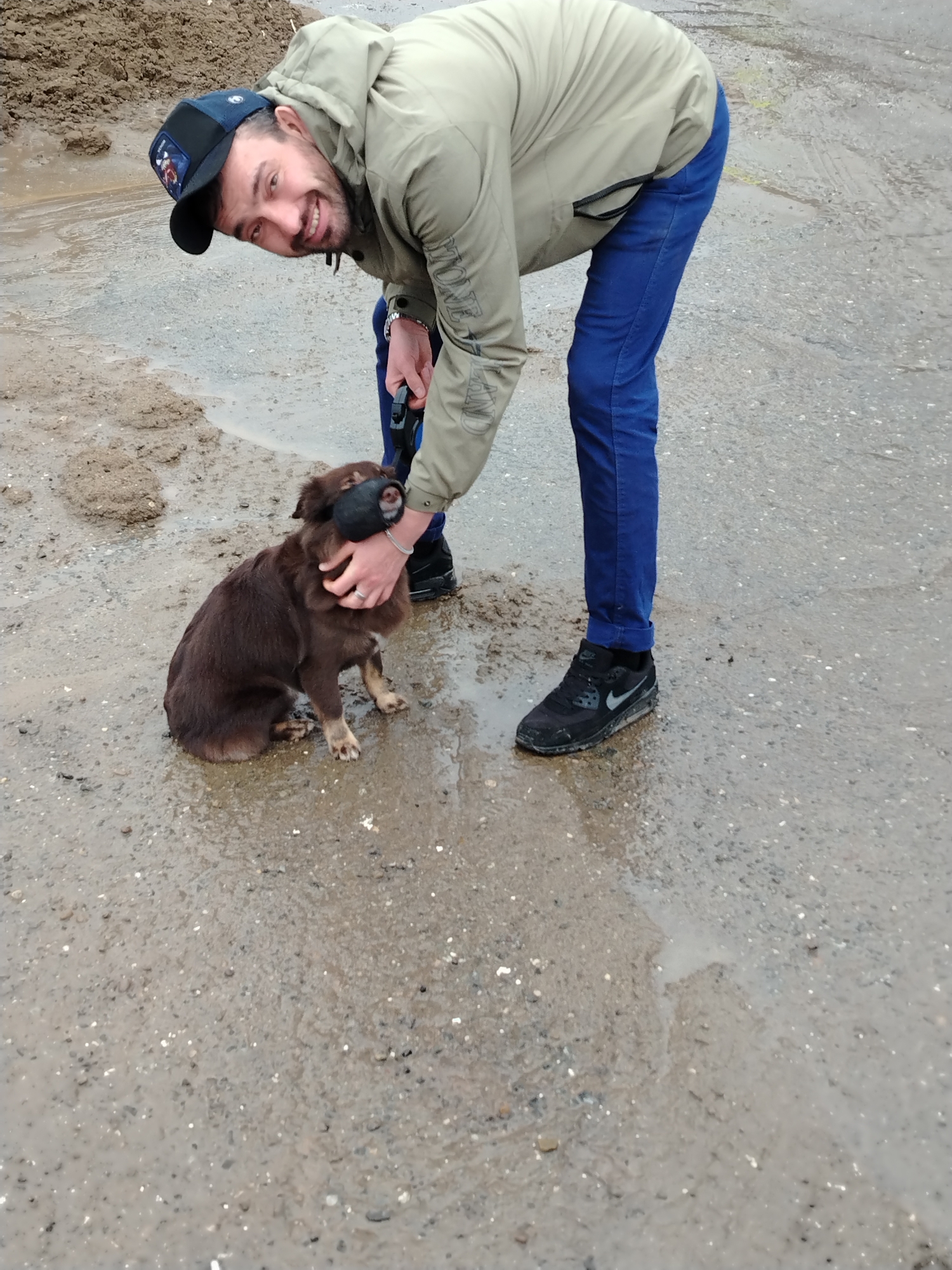 A medium outdoor shot depicts a man in a light green jacket and blue jeans petting a brown dog with a muzzle in a muddy puddle. The man is wearing a black cap and black Nike sneakers. He is bent over, smiling, with his right hand petting the dog’s head and his left hand holding the dog’s leash. The dog is sitting in the puddle, looking forward. The ground is muddy, and there is a small pile of dirt in the background. The location is in the city of Большая Уса, Russia.