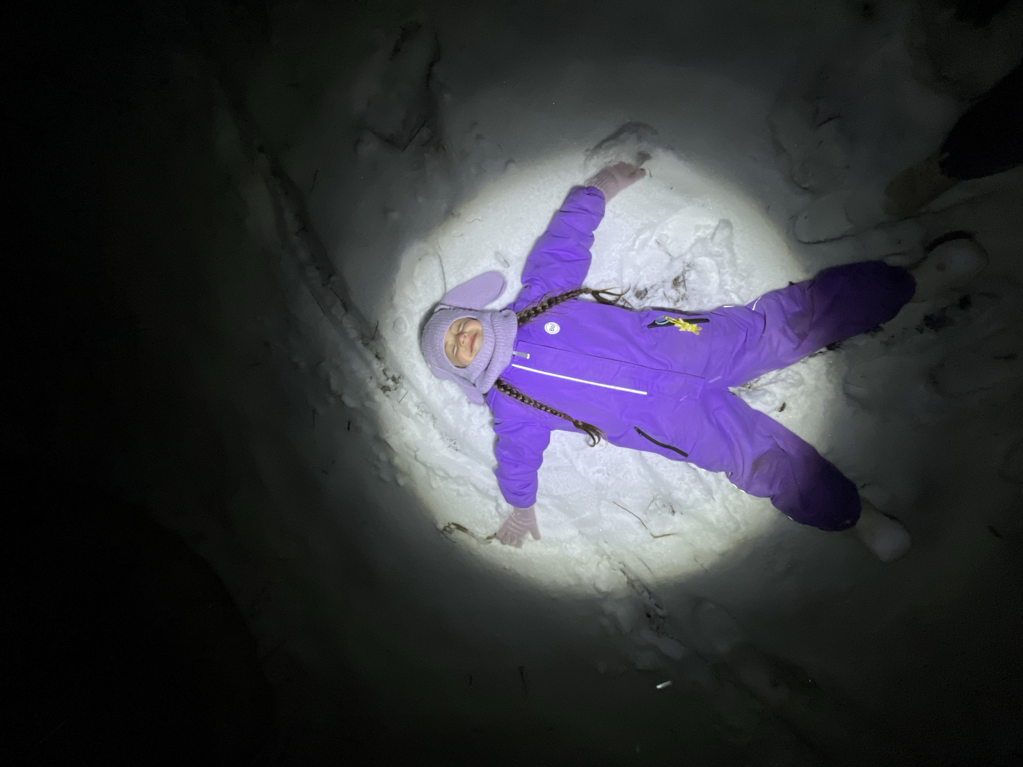 A young girl in a purple snowsuit and bunny-eared hat lies in the snow, making a snow angel. The surrounding environment is dark, with snow illuminated by a concentrated light source. The location, Pozim', Russia, is unconfirmed but assumed to be outdoors. Another individual is standing nearby, with their lower legs and feet visible at the right side of the frame.