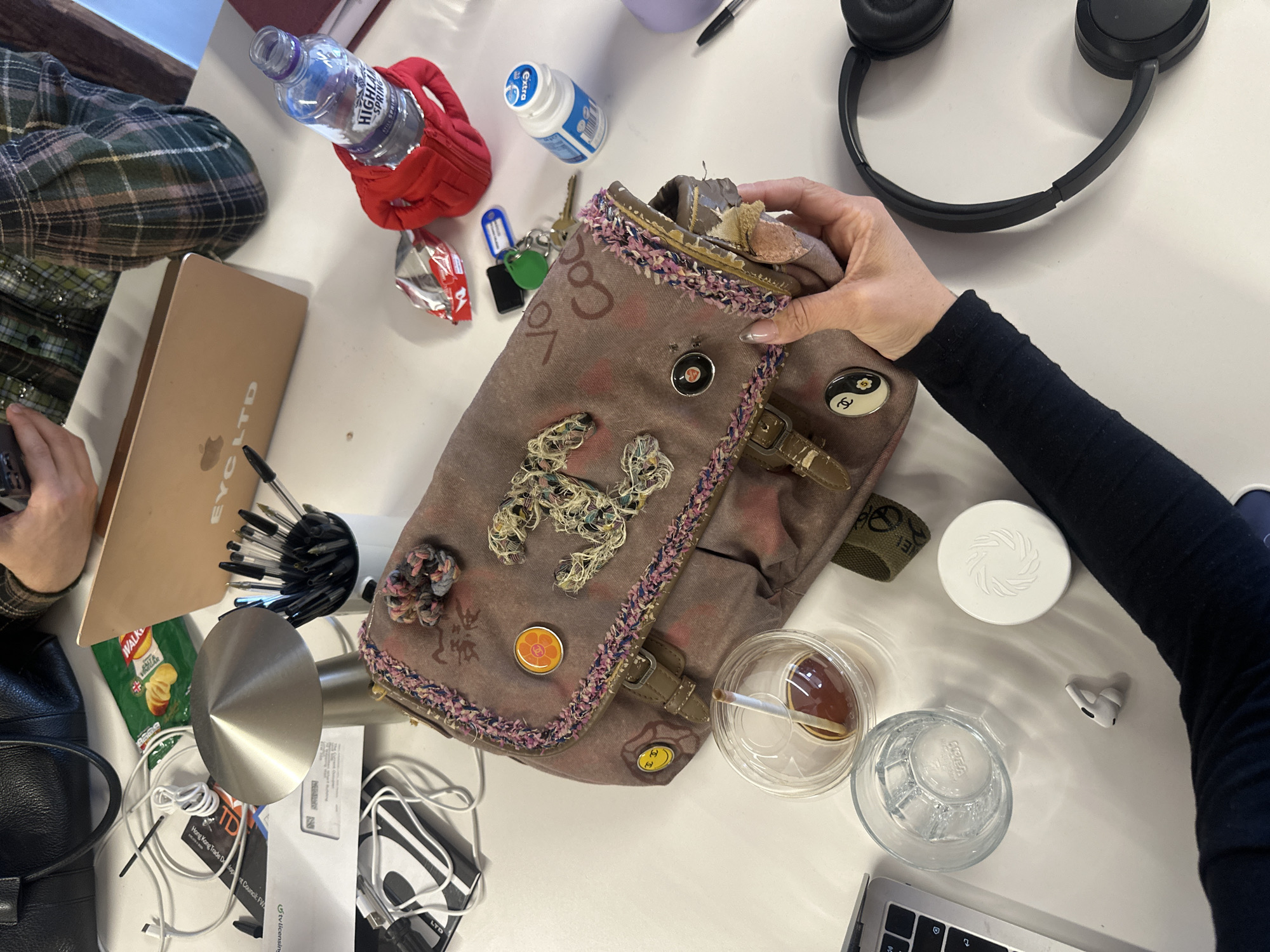 The image captures an overhead view of a cluttered desk or table, likely in an indoor office or workspace setting, possibly in London, United Kingdom. Two individuals are partially visible, with hands indicating their presence. On the left, a hand holds a dark object near a gold-colored MacBook laptop, while another person's right hand, with manicured nails, is seen reaching into or holding a distinctive fabric shoulder bag in the center.
The table is densely covered with a variety of personal and work-related items. Objects include a "HIGHLAND SPRING" water bottle in a red holder, a pack of "extra" gum, and a set of keys near the top center. Black over-ear headphones are placed near the upper right. The central fabric bag features intricate embellishments, including embroidered elements resembling a stylized "Chanel" logo, a tweed-like trim along its flap, and decorative pins such as a Yin-Yang symbol and a "Chanel" logo.
Other items visible on the table include a black leather bag, a pen holder filled with pens, various charging cables, and documents. A packet of "WALKERS" salt & vinegar crisps and a metallic conical object are near the left side. Towards the right, there are two clear plastic cups (one containing a beverage, possibly iced coffee, and another empty glass), a white round container with a spiral logo, and a single white earbud. Visible text on documents includes "Hong Kong Trade Development Council: FV" and "tv licensing." The scene suggests an active, informal working environment.