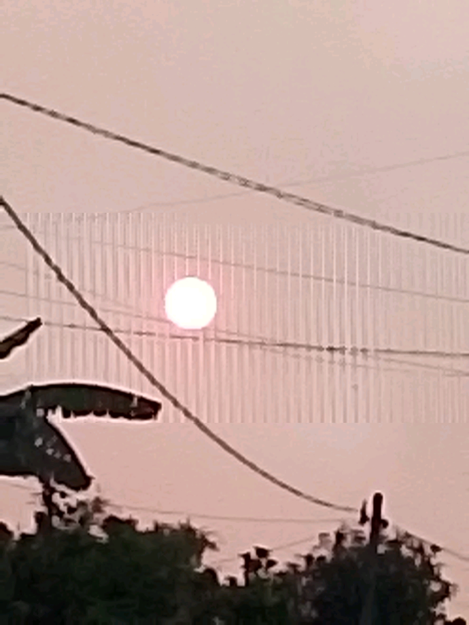 The image depicts a sky with a circular, luminous object, likely the sun, partially obscured by power lines. The sky is a pale pinkish-orange hue, suggesting either sunrise or sunset. In the foreground, the silhouettes of trees and what appears to be a structure with an angular, dark shape are visible. The environment appears to be outdoor, possibly in a semi-rural or suburban area given the presence of power lines. The location is specified as Bulurejo, Indonesia. The overall scene is somewhat hazy and lacks sharp detail due to the image quality.