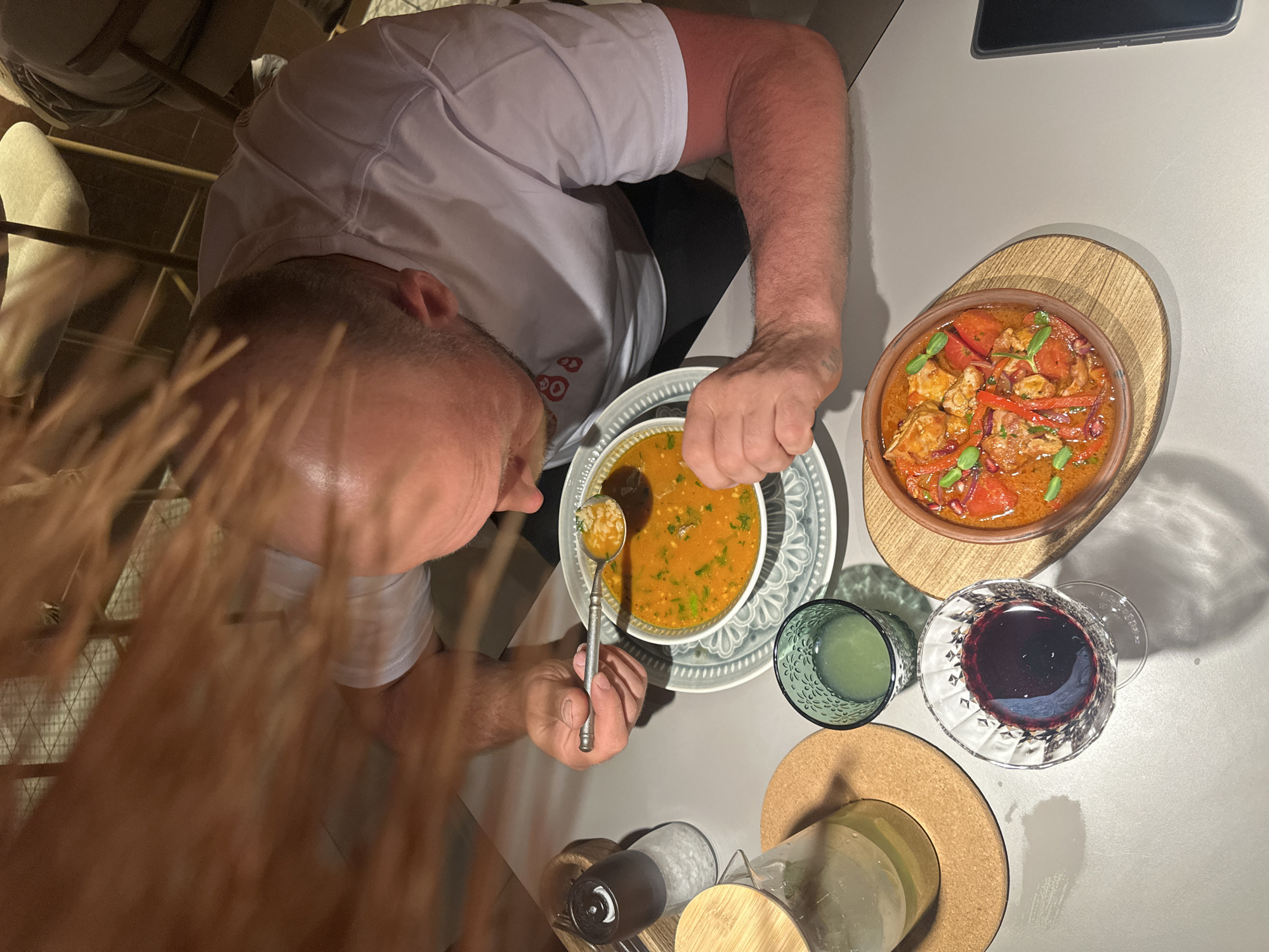 An overhead shot captures a man dining at a table. The man, wearing a white shirt, is in the process of eating soup from a white and blue patterned bowl with a silver spoon. Next to his soup is another dish served in a terracotta bowl placed on a wooden base, and a glass of red wine, alongside a green-tinted glass. Additional food items are present on the table near the condiments in the lower center of the image. The scene is situated at the location of Chaikovsky, Russia.