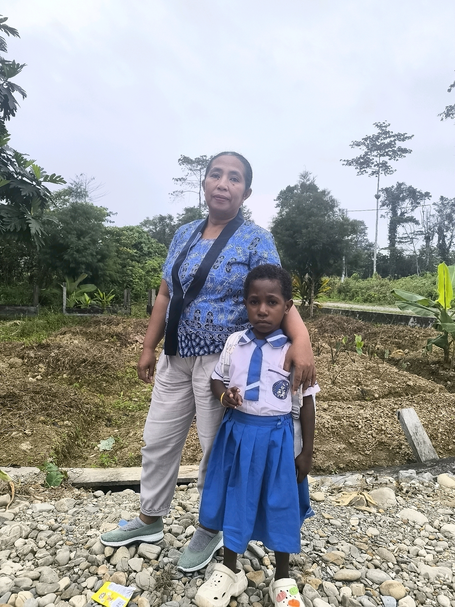 An adult woman and a young child stand outdoors in what appears to be a rural or semi-rural area, possibly on the outskirts of Timika, Indonesia. The ground is covered with rocks and dry earth, with some patchy vegetation. In the background, there are trees, some of which are quite tall, and a cloudy sky.
The woman is wearing a blue patterned shirt and light-colored pants. She has her arm around the child. The child, who appears to be a student, is wearing a white collared shirt with a blue tie and a blue pleated skirt. Both are looking towards the camera. The child's expression is neutral.
The overall scene suggests a moment captured between the woman and the child in a somewhat undeveloped environment.