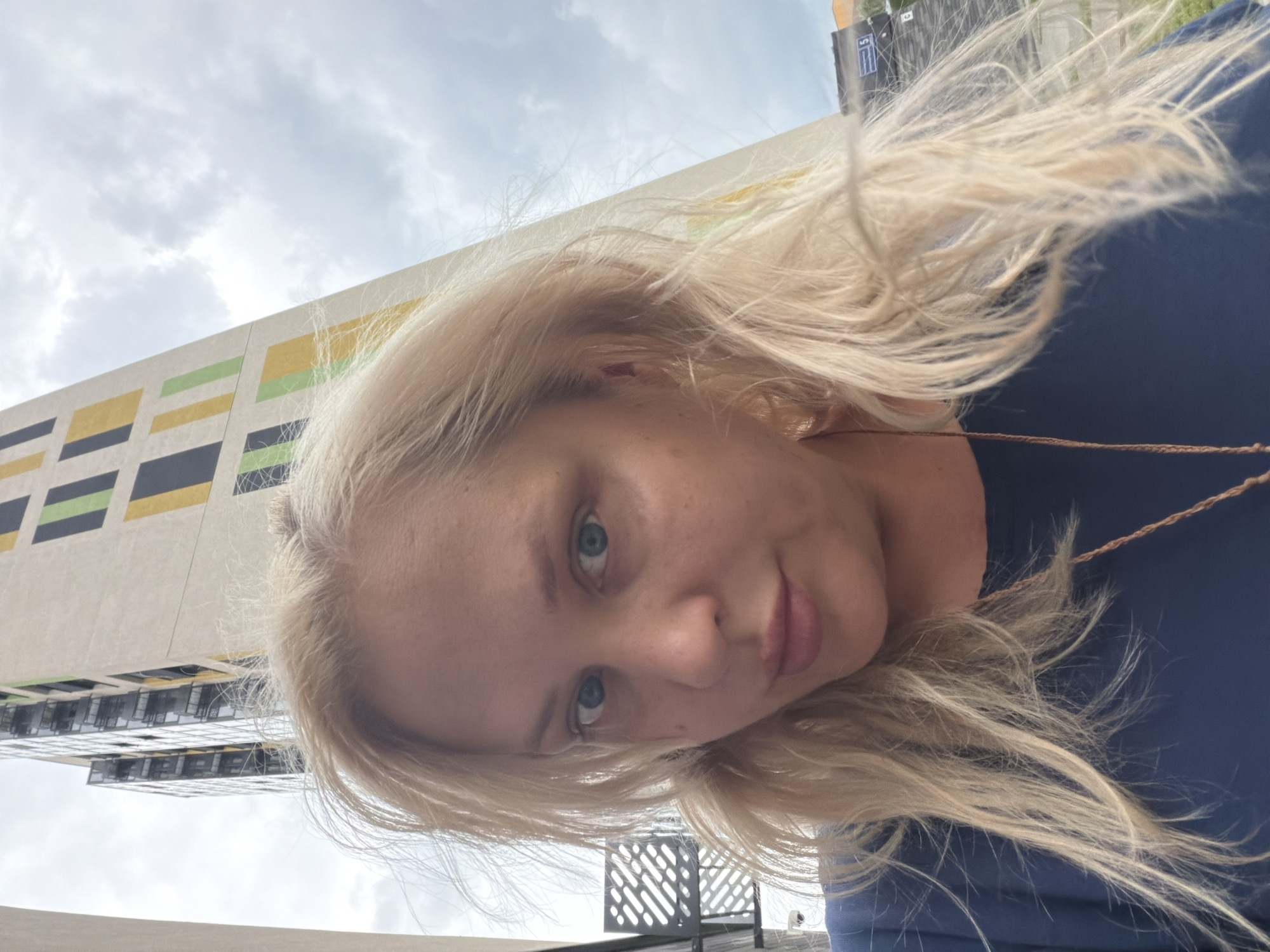 A blonde woman with blue eyes and fair skin poses for a selfie in front of a tall, modern building with a distinctive facade of colored rectangular panels. The building, located in Kazan, Russia, has a beige base with vertical stripes of black, yellow, and green. The sky is overcast with clouds. The woman is wearing a dark blue shirt and a thin brown cord necklace. Her blonde hair is windswept. To the left of the woman and behind her, a portion of a dark metal structure with a patterned surface is visible. The image is shot from a slightly upward angle.