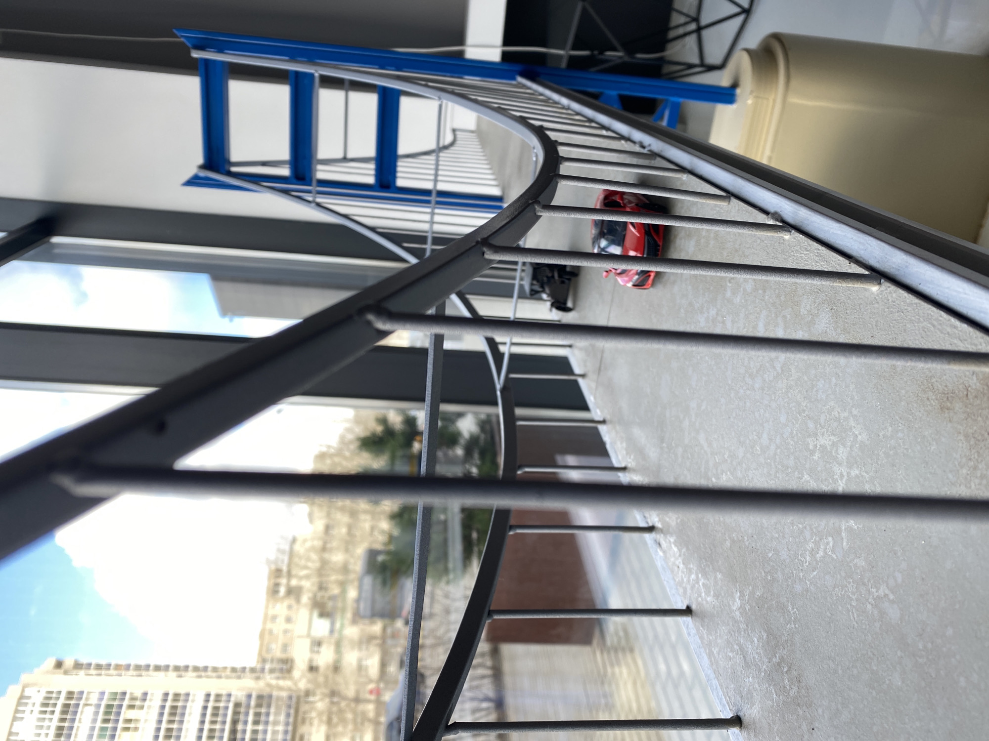 The image shows a spiral staircase viewed from a high angle, likely from the next level up. The railing is gray and has vertical bars. Outside the window, a cityscape is visible, with a building on the left side of the image. On the stair landing, there is a red toy car and a small, black object resembling a camera or microphone. The location might be Yekaterinburg, Russia, although there is no definitive information in the image to confirm this. There are no people visible in the frame. A blue object, possibly a ladder, is visible further up the staircase. A large, cylindrical beige object is partially visible in the upper right of the frame.