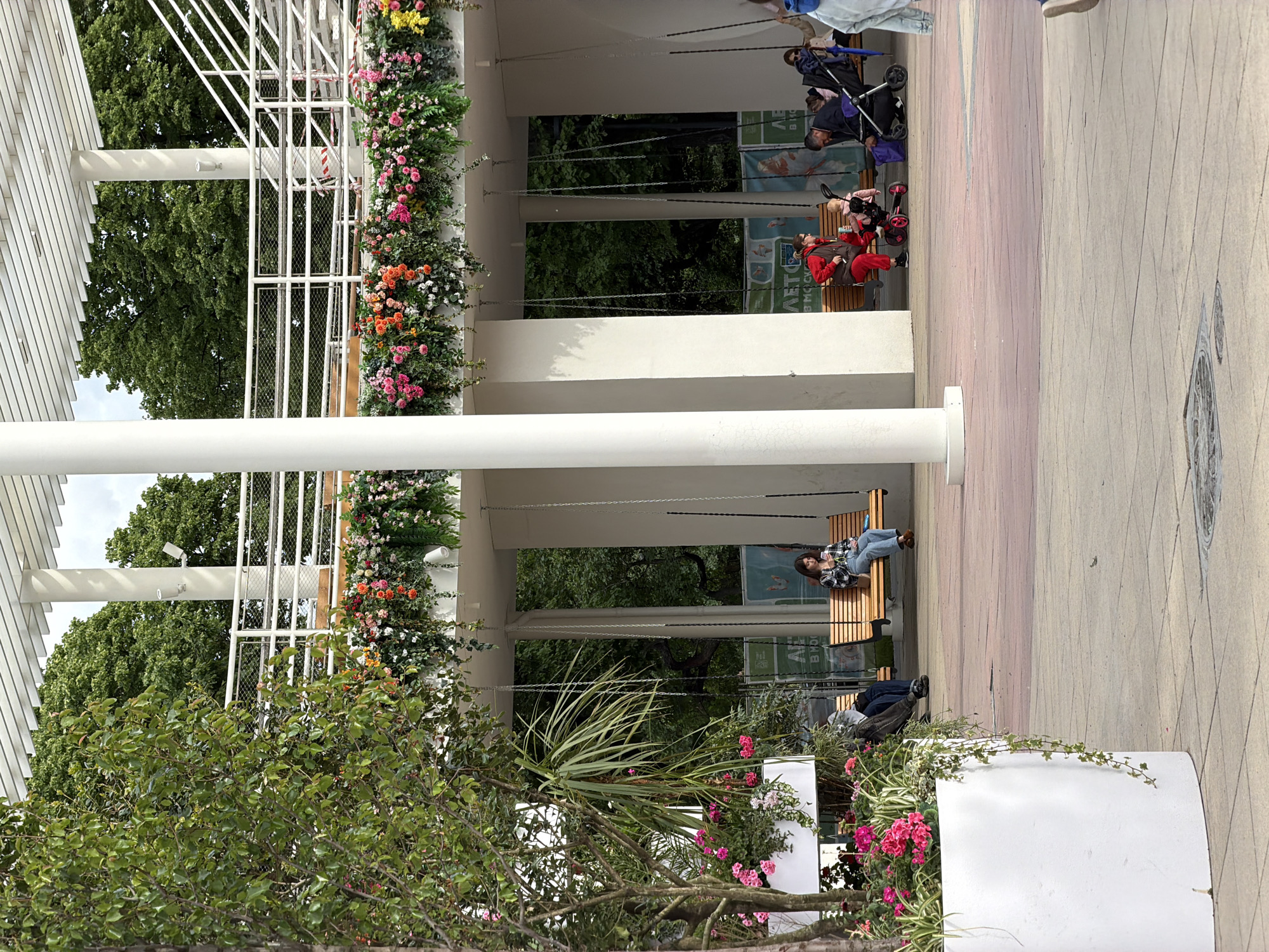 The image depicts an outdoor space, possibly a park or public area, in Moscow, Russia. The scene features covered swings suspended from a structure adorned with flowering plants. People are utilizing the swings, including an individual lying down and others sitting. Additional people are present, some with strollers, suggesting a family-friendly environment. The ground is paved, and trees are visible in the background, contributing to the park-like setting. Flower beds with pink blooms are also present in the foreground.