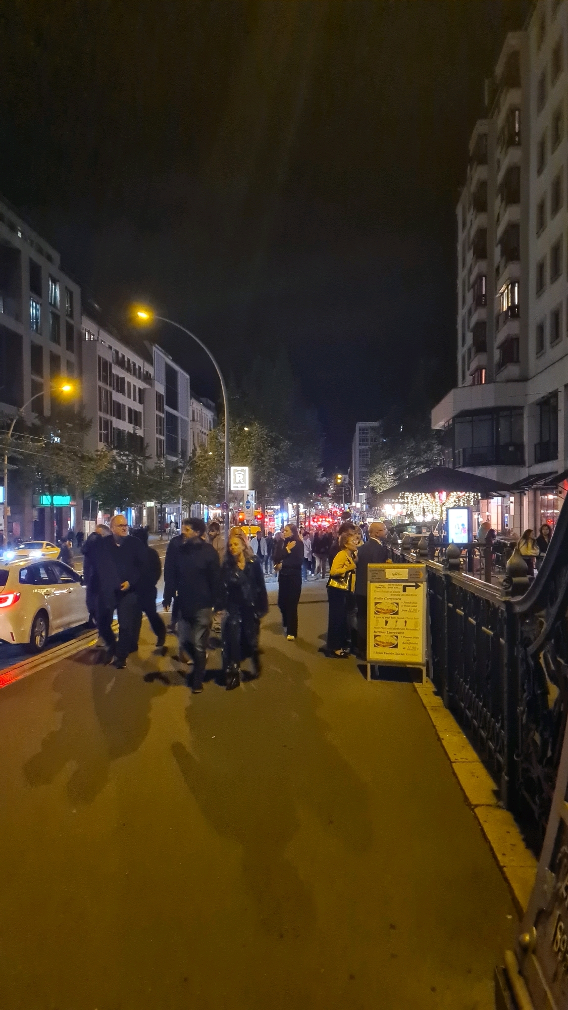 The image depicts a street scene at night in Berlin, Germany. A crowd of people are walking along a sidewalk and across a street. Several buildings, some appearing to be apartment buildings and others commercial establishments, line the street. Streetlights illuminate the scene. A white car is parked on the street. A sign is visible on the sidewalk with text in German. The overall atmosphere is one of urban nightlife.