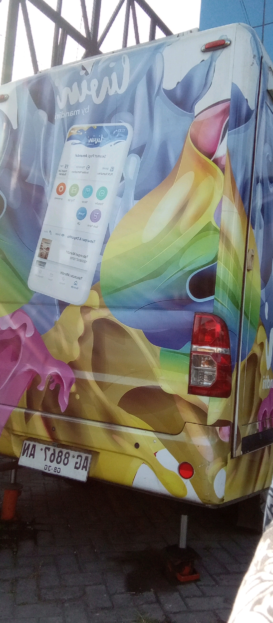 A white van with a colorful, abstract design is parked on a cobblestone surface, likely a street or parking lot. The design features flowing shapes in shades of yellow, pink, blue, and green, with a smartphone graphic overlayed on the left side. The van has a visible license plate with the characters "MA - 1888 DA" and the text "08-80" below it. A red taillight is visible on the right rear of the van. A person's arm, wearing a patterned sleeve, is partially visible in the lower right corner of the image. The background shows a metal structure, possibly part of a building or bridge. The scene appears to be outdoors in daylight.