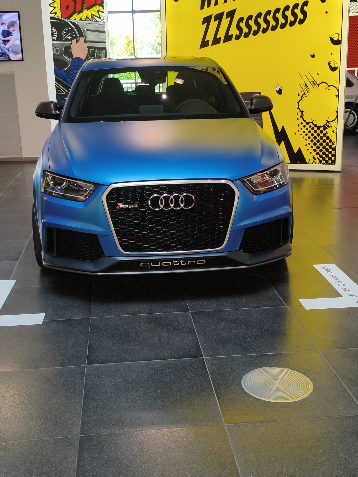 The image shows the front of a matte blue Audi RS Q3 concept car parked indoors on a tiled floor. The car's grille features the Audi rings logo and "RS Q3" badging, with "quattro" written below it. In the background, a large yellow wall with black comic-style graphics and the text "ZZZssssssss" is visible. To the left, a portion of another graphic with a steering wheel and hand is seen, along with a screen displaying people. The car is positioned in what appears to be a showroom or exhibition space, with white floor markings indicating a path or zone. The environment suggests a modern automotive display, potentially at the Audi Forum in Ingolstadt, Germany, a known location for such exhibitions and brand experiences. There are no people directly interacting with the car. The lighting is even, illuminating the car and the background graphics.