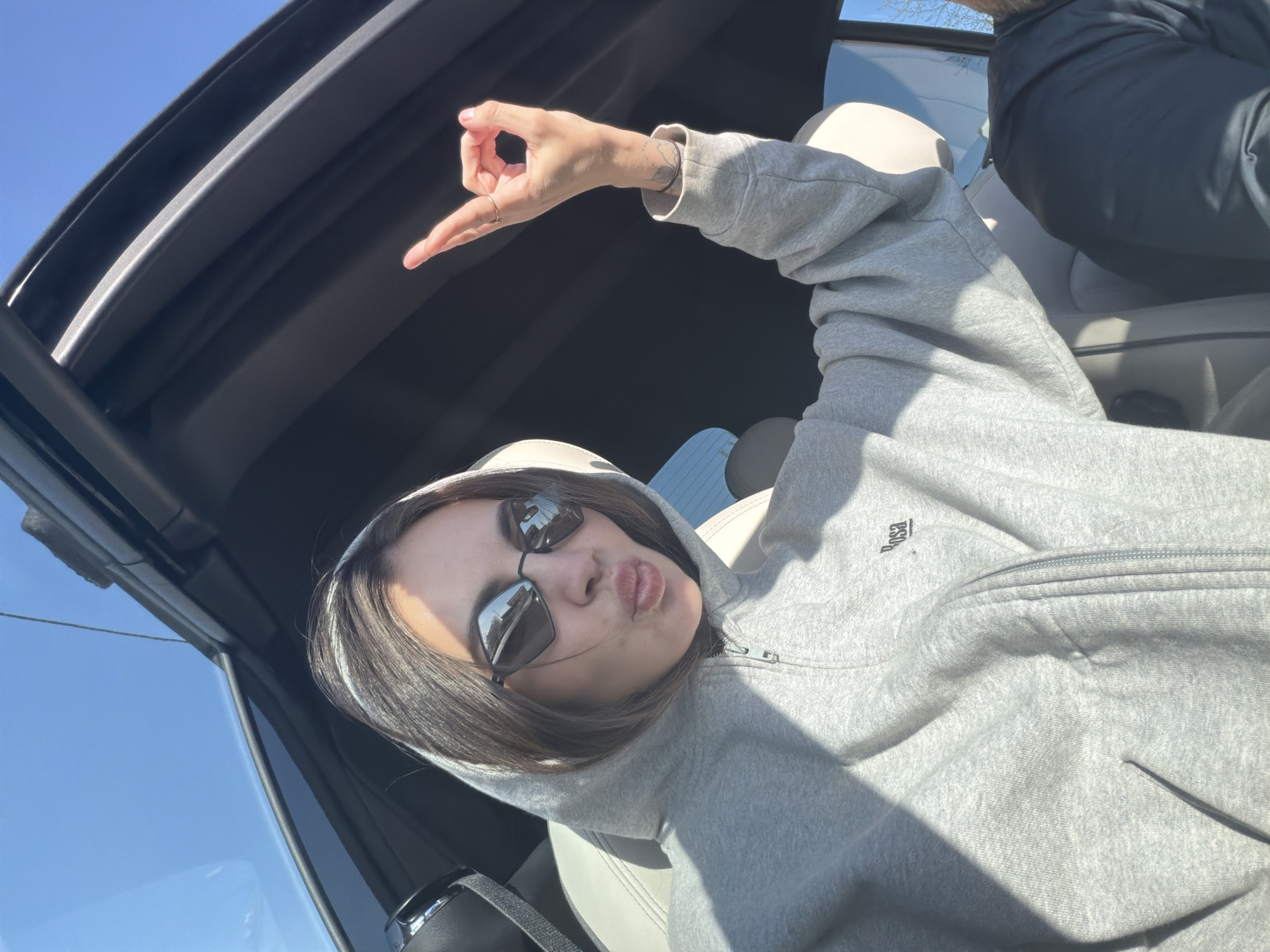 A person wearing sunglasses and a gray hooded sweatshirt is inside a car, possibly a convertible, making a hand gesture with their right hand. The interior of the car features light-colored seats. Outside the car is bright, implying a sunny day. The image does not provide any information that allows determining that the location is Zheleznogorsk, Russia.
