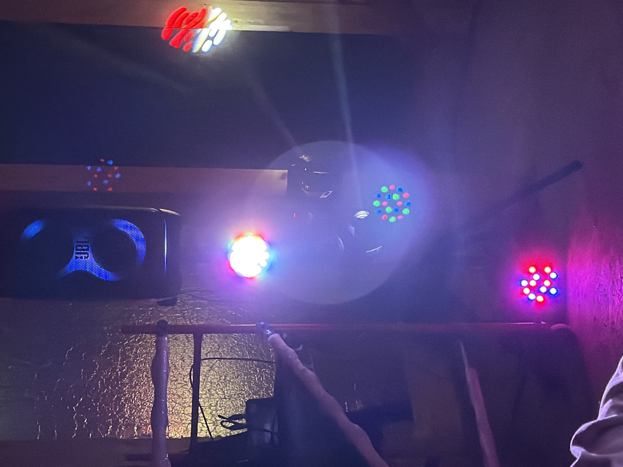 The image shows a dimly lit interior space with several light sources. There is a black JBL speaker with blue accents visible on the left side of the image. Multiple disco-style LED lights are visible in various colors and patterns, positioned around the room and on the ceiling. The light creates a diffused and bright effect, obscuring details in the background. There is a metallic-looking textured surface behind the speaker, and a red railing or support structure runs horizontally across the middle of the frame. A portion of a person's arm and shoulder are visible in the bottom right corner. The exact location within the city of Chaikovsky, Russia is not discernible from the image.