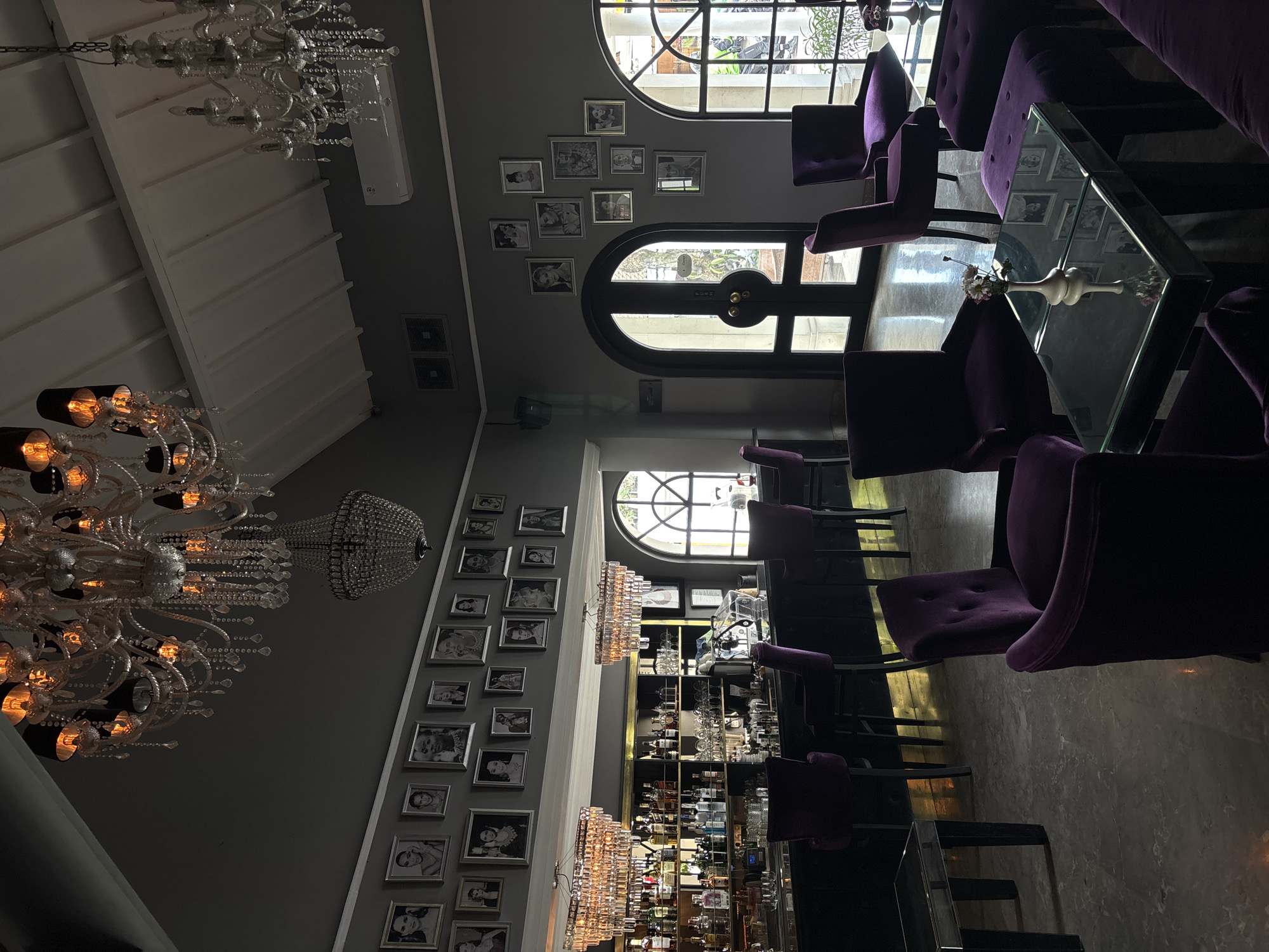 The image shows the interior of a dimly lit, upscale bar with a focus on dark purple velvet seating and opulent chandeliers. The bar itself is well-stocked with bottles of alcohol and glassware, suggesting it is operational. Portraits of various individuals are displayed on the grey walls, adding a decorative element to the space. Several chandeliers, one large and ornate and another crystal, hang from the ceiling, casting a warm glow. The furniture consists of plush purple chairs, stools, and ottomans, arranged around glass-topped tables. Large arched windows offer glimpses of the outdoors, and an air conditioning unit is visible on the wall. The flooring appears to be polished stone. The overall ambiance is sophisticated and luxurious, indicative of a high-end establishment in North Kuta, Indonesia. There are no people visible in the image.