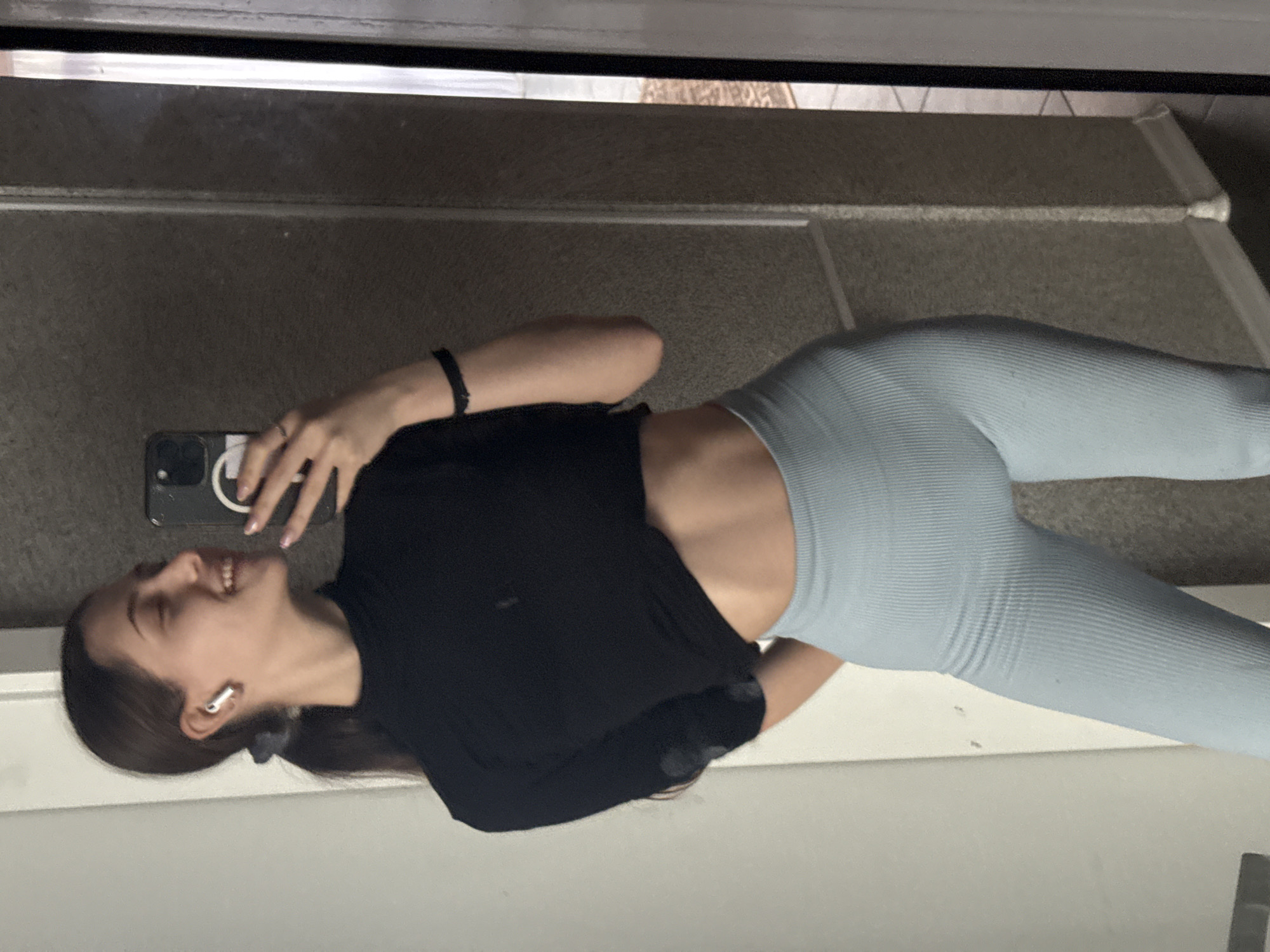 A woman is positioned at an angle, holding a phone in her right hand, appearing to take a selfie. She is wearing a black t-shirt and light blue leggings. An Apple AirPod is visible in her left ear. She has dark hair pulled back in a ponytail and appears to be smiling. The background reveals a neutral-toned floor, potentially a tile or concrete surface with a metallic threshold and a white painted surface. There is no information available to deduce the city of Sofia, Bulgaria, from the image.