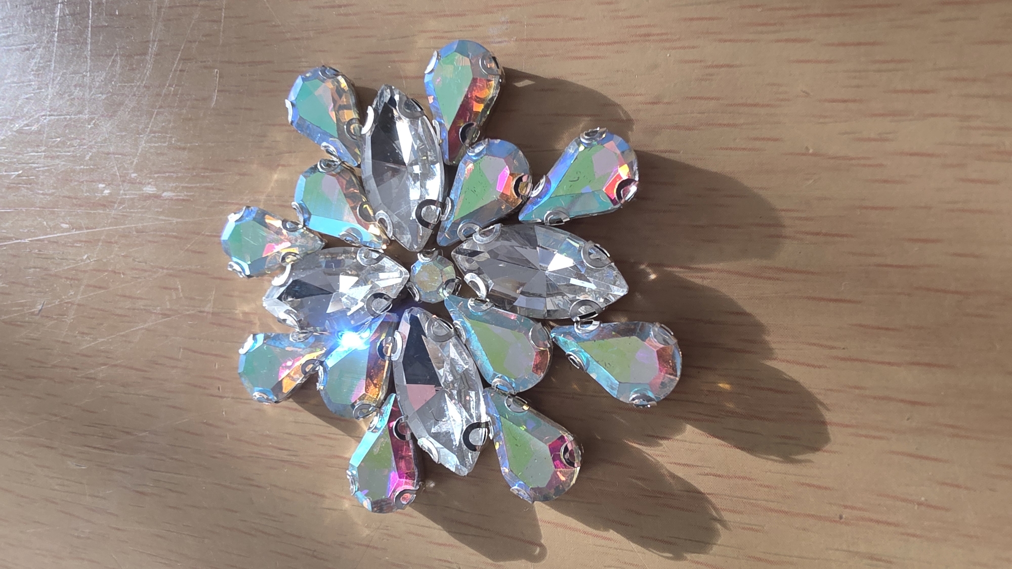 The image shows a decorative arrangement of iridescent teardrop and oval-shaped rhinestones on a light brown wooden surface. The rhinestones are arranged in a roughly circular pattern, resembling a flower or starburst. The light source appears to be coming from the upper left, casting shadows to the right of the arrangement. The scene provides no evidence to determine a location in Blagoevgrad, Bulgaria.