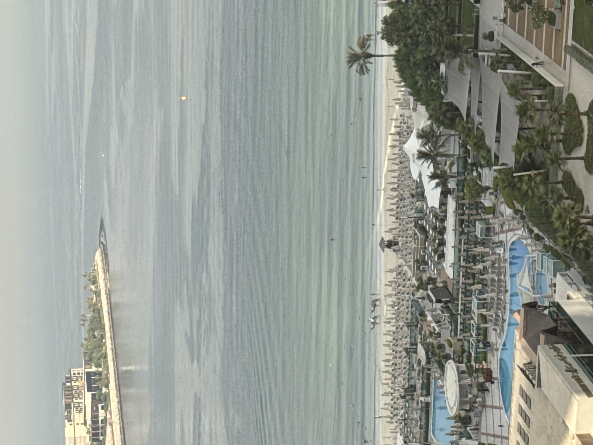 This image captures a panoramic view of a bustling beachfront resort in Dubai, United Arab Emirates. The scene is dominated by the vast expanse of the ocean, its waters a tranquil gradient of blues and greens, marked by subtle ripples and a few distant buoys. On the shore, a wide stretch of beach is meticulously lined with numerous white umbrellas and lounge chairs, indicative of a popular tourist destination.
In the foreground, a complex of resort facilities unfolds, featuring several swimming pools of varying sizes and shapes, some shimmering blue, others with intricate water features. Cabanas and shaded seating areas are scattered throughout the resort grounds, interspersed with lush green palm trees and manicured landscaping. Buildings with modern architecture, some resembling luxury hotels or residential complexes, are visible at the edges of the resort and in the distance.
Notable elements within the scene include two camels with riders on the beach, a typical attraction for tourists in Dubai, suggesting leisure activities are underway. A few individuals are discernible on the beach and near the pools, engaged in various activities or simply relaxing. The overall impression is one of a vibrant and well-appointed resort, embodying the luxurious lifestyle often associated with Dubai. The clear, bright lighting suggests a daytime setting, likely during a warm season. The image is taken from an elevated vantage point, providing a comprehensive overview of the resort and its surrounding environment.