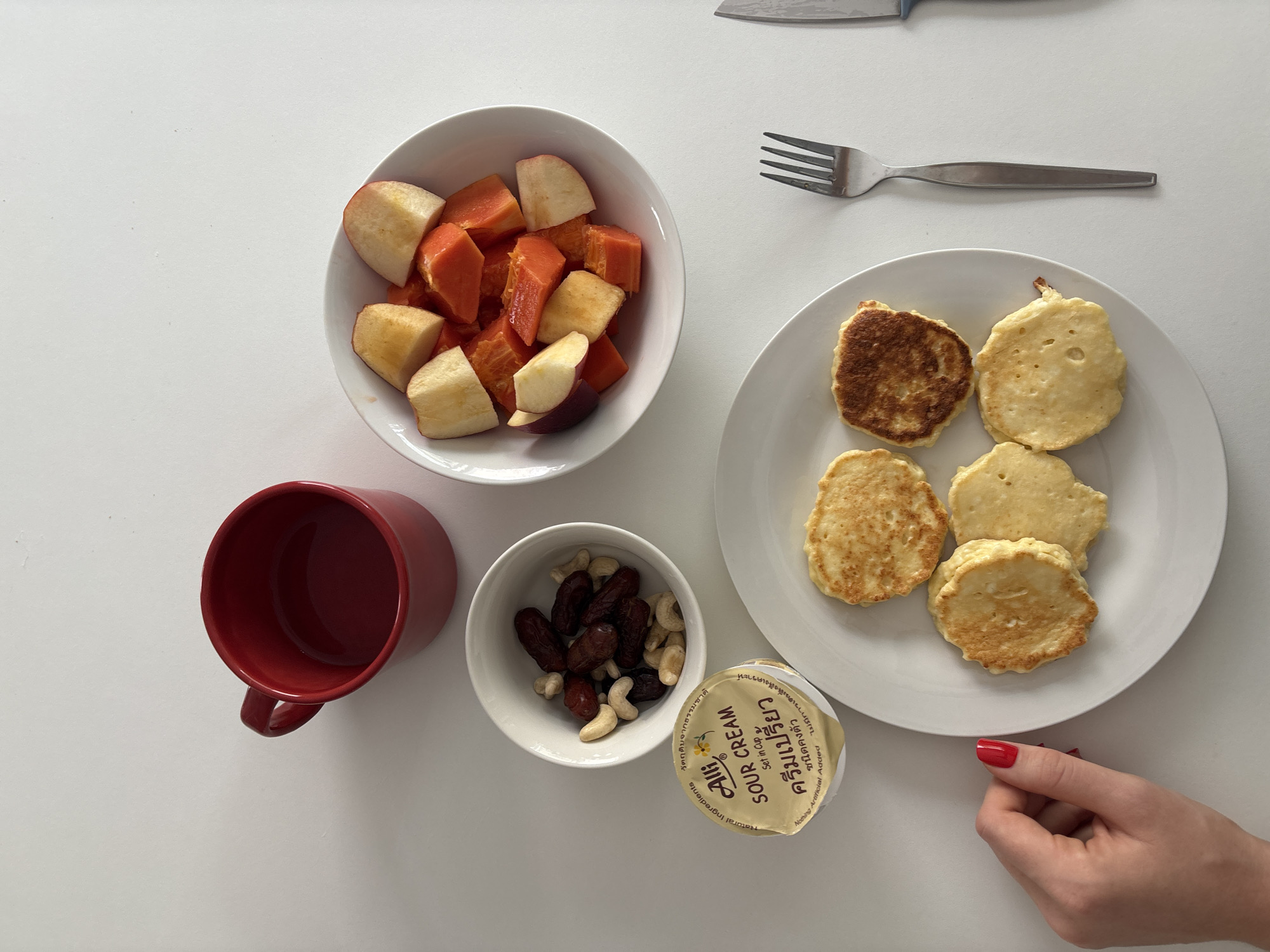 A flat lay image taken from above shows a breakfast spread on a white surface. The scene is likely from a home kitchen in Ko Kaeo, Thailand, as indicated by the Thai writing on the sour cream container.
In the upper left is a red ceramic mug. To its right, a white bowl contains chopped fruit, primarily papaya and apple. Below this, a small white bowl holds dates and cashews. A single-serving container of "Alli Sour Cream" is positioned in front of the bowl of nuts and dates.
To the right of the sour cream and nuts is a white plate with four small, round pancakes, some with golden-brown edges. A hand with bright red nail polish is reaching into the frame from the lower left, near the pancakes. To the right of the plate, a silver fork and knife are laid out. The white surface appears to be a tabletop.