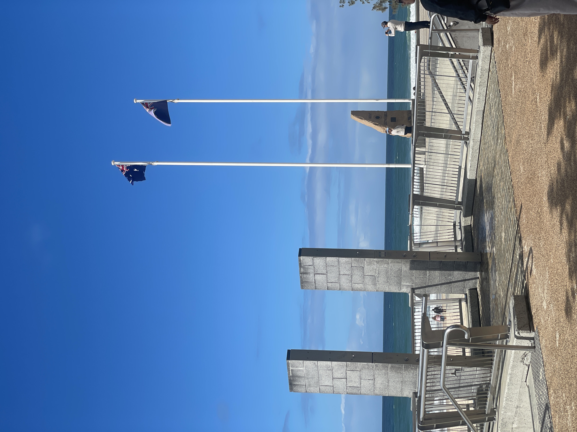 The image captures a section of the Gold Coast, Australia's coastline, near a memorial. Two large, grey, rectangular stone pillars stand prominently in the foreground, with metal railings and a paved pathway leading towards the beach. Beyond these structures, the ocean stretches out under a clear blue sky, with gentle waves breaking on the sandy shore. Two Australian flags fly from tall white poles, one on the left with its flag partially obscured by wind, and another on the right. In the middle ground, a rough, angular stone monument inscribed with "ANZAC" and "LEST WE FORGET" is visible between the flagpoles. A few people are present on the beach and near the memorial area. One individual is walking on the beach with another person, while a man in the mid-right of the frame is seen taking a photograph with a phone, and another person stands near the monument. The overall scene depicts a bright, sunny day at a coastal memorial site.