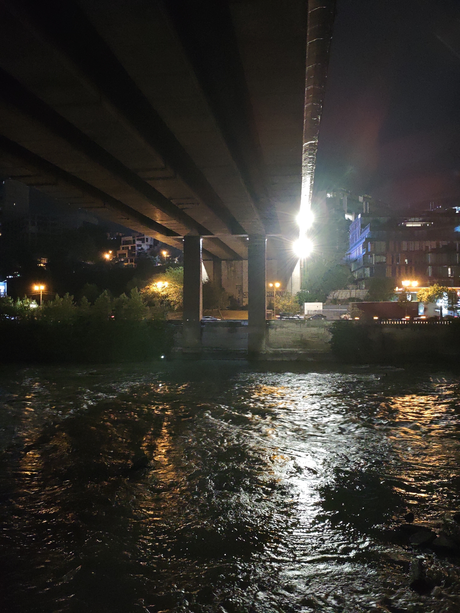 The image captures a nighttime view of the Mtkvari River in Tbilisi, Georgia, flowing beneath a large concrete overpass. The water is dark and choppy, reflecting the bright yellow streetlights and building lights from the riverbank. The overpass itself is a dominant feature, with its thick concrete beams casting deep shadows. A large concrete pillar supporting the overpass is visible on the right side. The riverbank on the right is lined with trees and buildings, some of which are illuminated by artificial lights, suggesting an urban environment. A truck with its lights on is visible on the road adjacent to the river. The overall atmosphere is dimly lit and somewhat industrial, illuminated by the scattered urban lights.