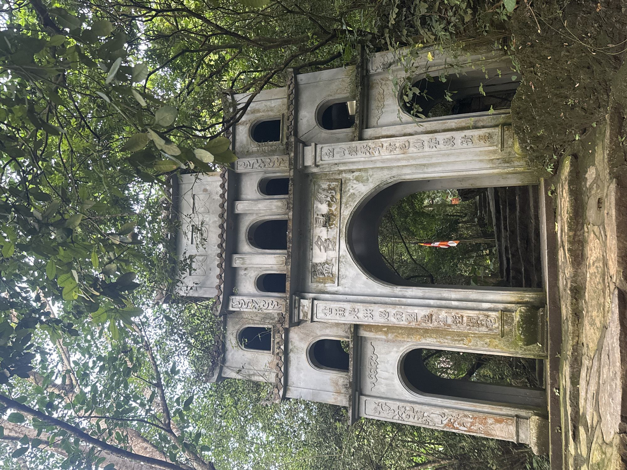 The image shows a weathered, ornate gateway structure in a lush, green forest setting. The gateway, made of gray stone, has multiple arched openings and decorative carvings. It appears to be part of an ancient temple or monument. The structure is adorned with Chinese characters, and there is a flag visible through the central archway. Stone steps lead up to the gateway and continue beyond it into the forest. The overall scene suggests a historical and spiritual site located in a natural environment. The location is in Tây Hoa Lư Ward, Vietnam.