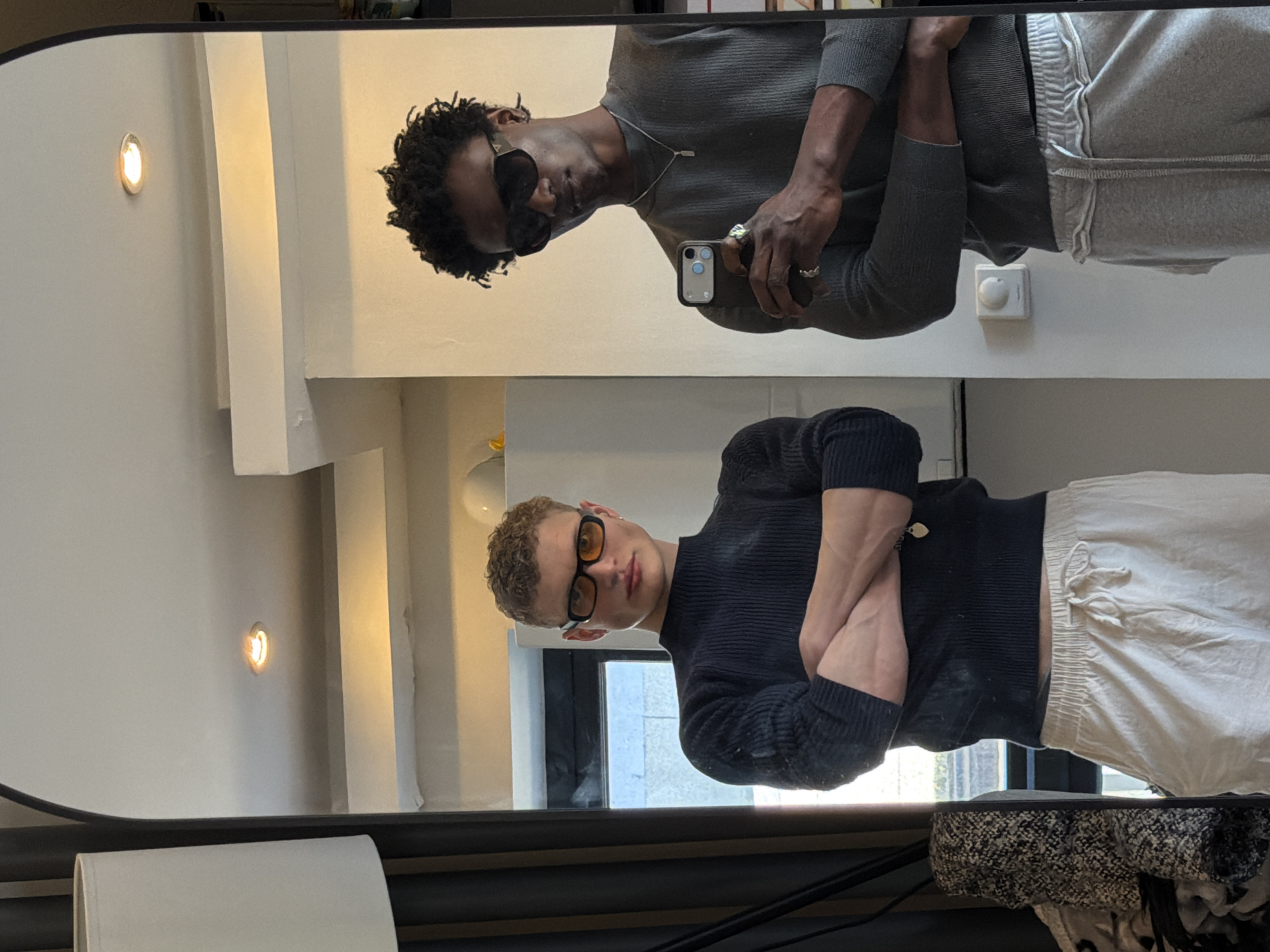 The image shows a mirror reflecting two individuals. The top reflection features a dark-skinned person with short, curly black hair, wearing sunglasses, a grey long-sleeved shirt, grey sweatpants, and a silver necklace. They are holding a smartphone. The bottom reflection is of a light-skinned person with short, curly blond hair, wearing yellow-tinted glasses, a navy long-sleeved shirt, and white sweatpants. Their arms are crossed. The background indicates an interior setting. Given the architectural style and lighting fixtures visible in the reflection, the location is likely an upscale residential or commercial space. While the precise location in London cannot be determined, the interior design suggests an affluent area within the city.
