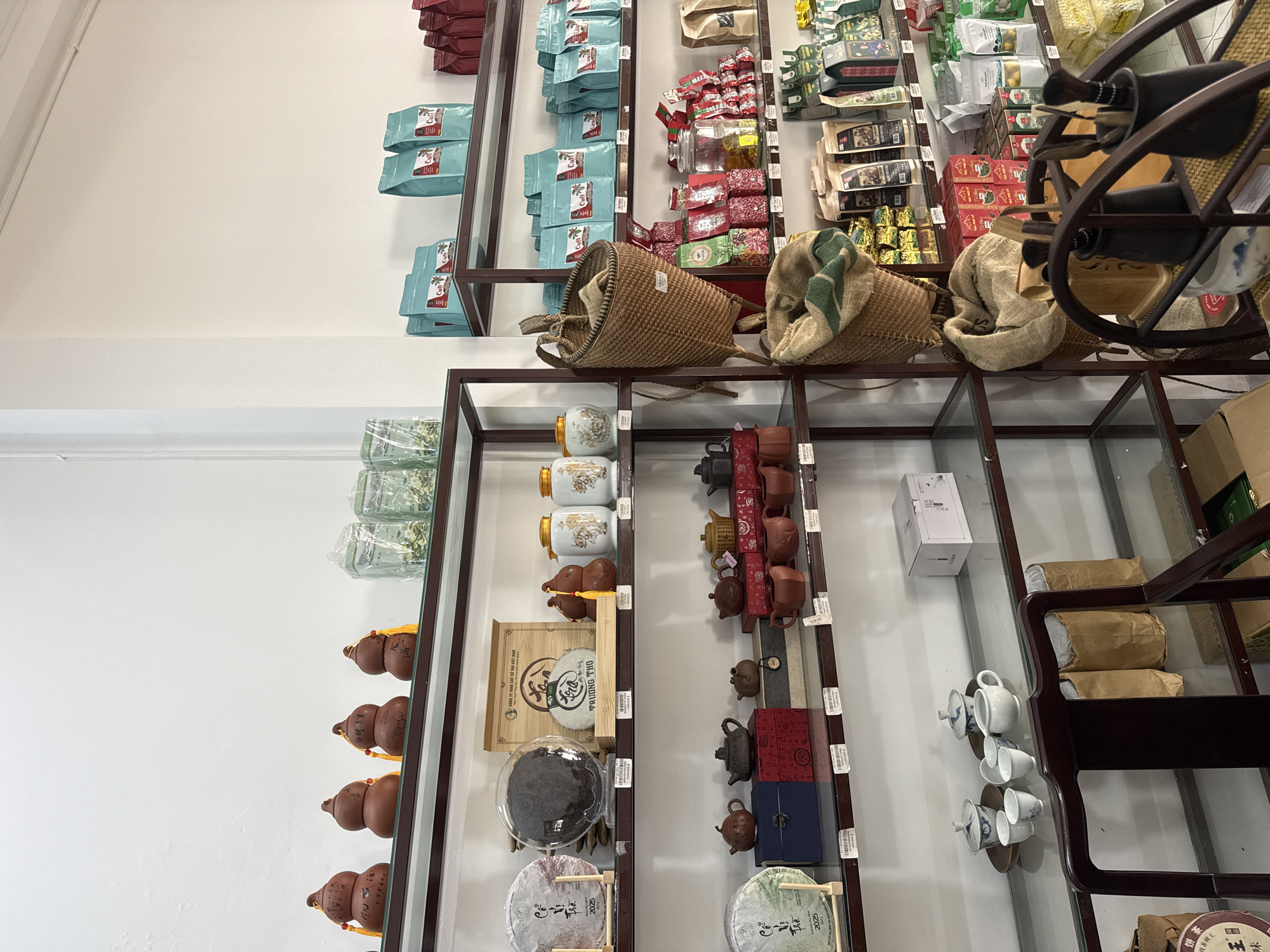 This image depicts the interior of a tea shop located in Bac Nha Trang Ward, Vietnam. The shop is filled with shelves displaying a variety of tea-related products. On the top shelf, several gourd-shaped ceramic items with tassels are visible, alongside glass jars containing tea leaves. The second shelf from the top features packaged tea, some in blue bags, and red packaged items. A woven basket is also present on this shelf. The third shelf displays more packaged tea in blue bags, as well as a glass jar filled with yellow spherical items and various red and white packages. Further down, the shelves are stocked with round cakes of compressed tea, ceramic teapots in different sizes and colors, and boxes of tea. On the bottom shelves, there are more packaged teas, some in colorful bags, and a set of white ceramic teacups and a teapot. A dark wooden chair is positioned in the foreground, with paper bags and a basket visible behind it. The walls are white, and the shelves are made of metal and glass. No people are visible in the image.
